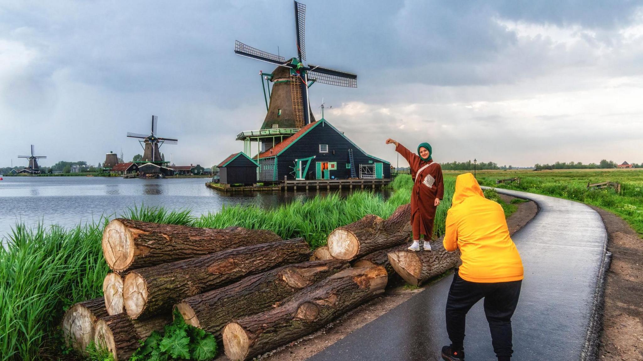 Woman posing in front of a windmill while someone in a yellow raincoat takes a picture 