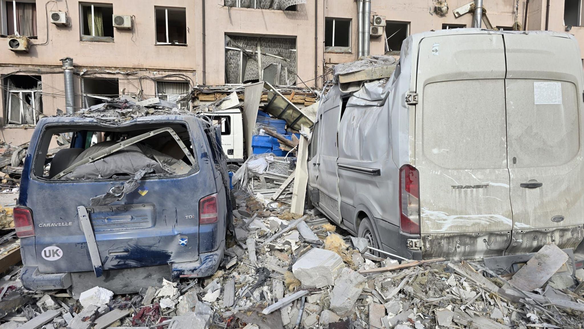 A close up of the damage to Karol's van. WIth the beige building hit by the missile in the background. All the glass from the windows is gone, from the building and the vans, they are covered and surrounded by rubble.