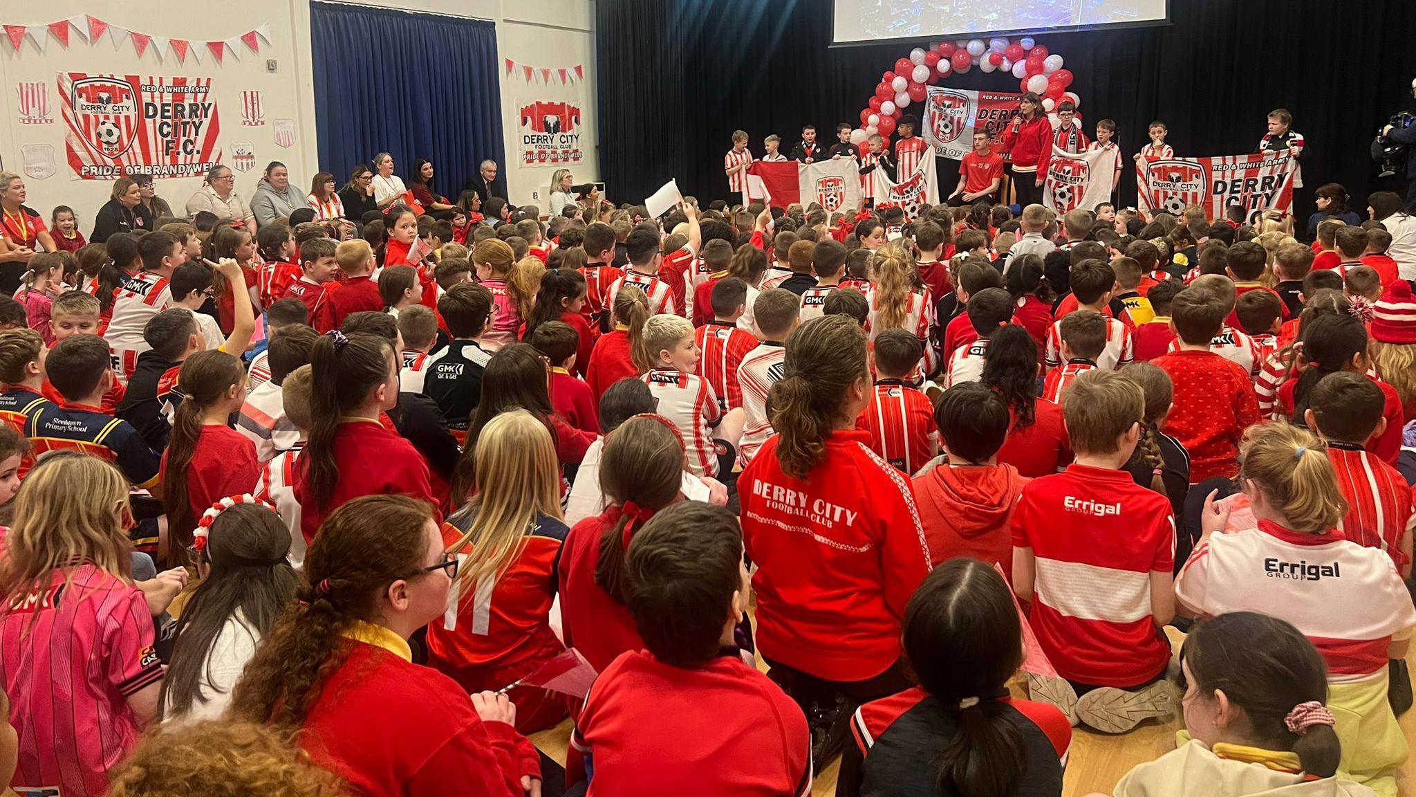 A wide shot of a school assembly hall filled with pupils wearing red and white Derry City and Derry GAA shirts - there are red and white balloons and Derry City banners on the walls