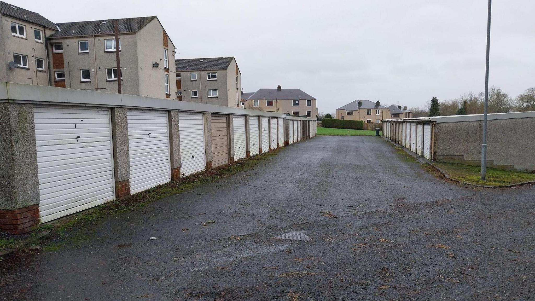 Two rows of stone garages with white doors. In the background are blocks of flats.