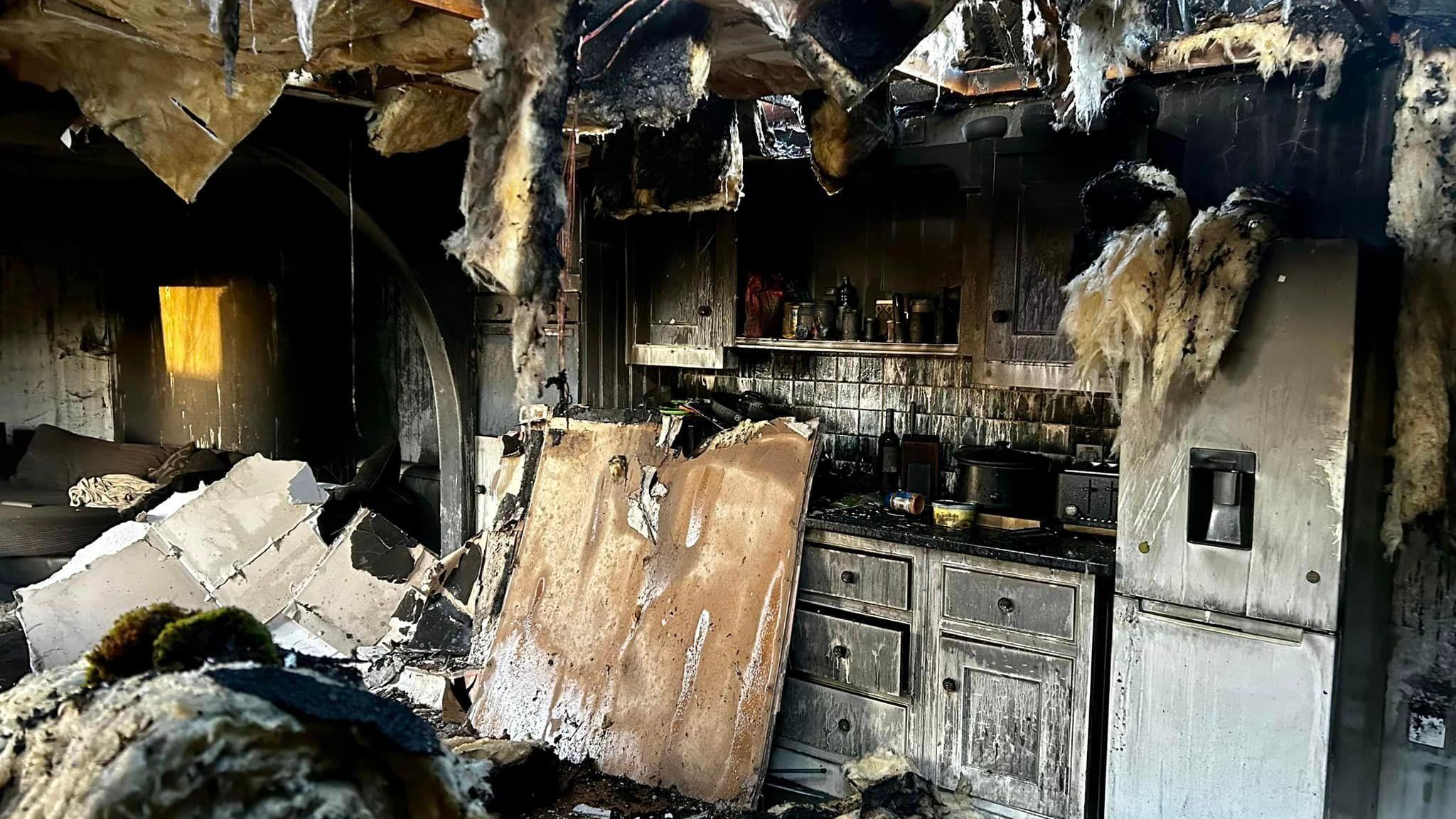 A photo of the inside of a property which has been severely damaged in a fire. Parts of the roof can be seen hanging down and black soot lines many of the surfaces and walls.