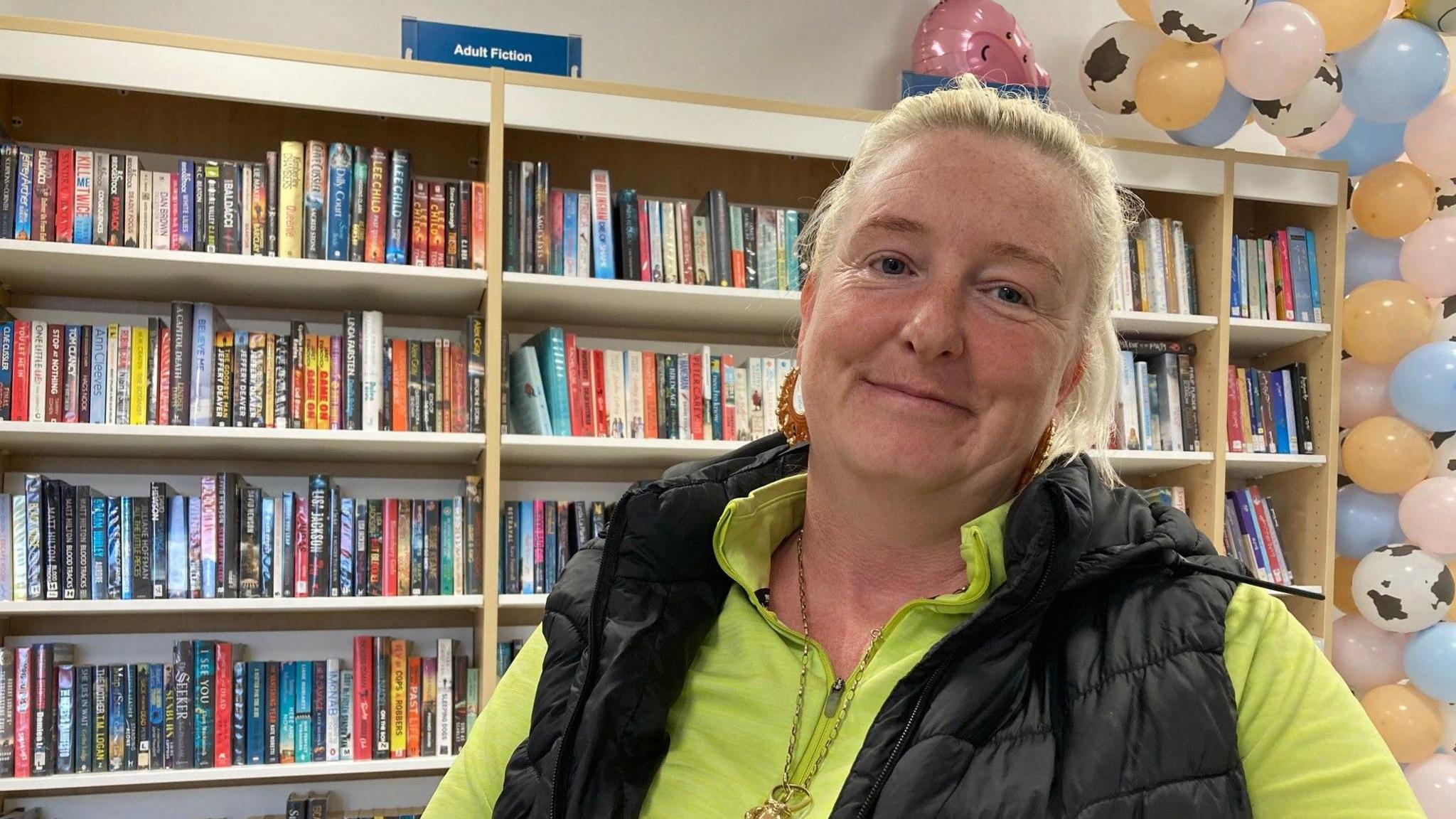 A woman with light blonde hair wearing a black gillet and a greentracksuit top underneath with rows of books on shelves in the background. 