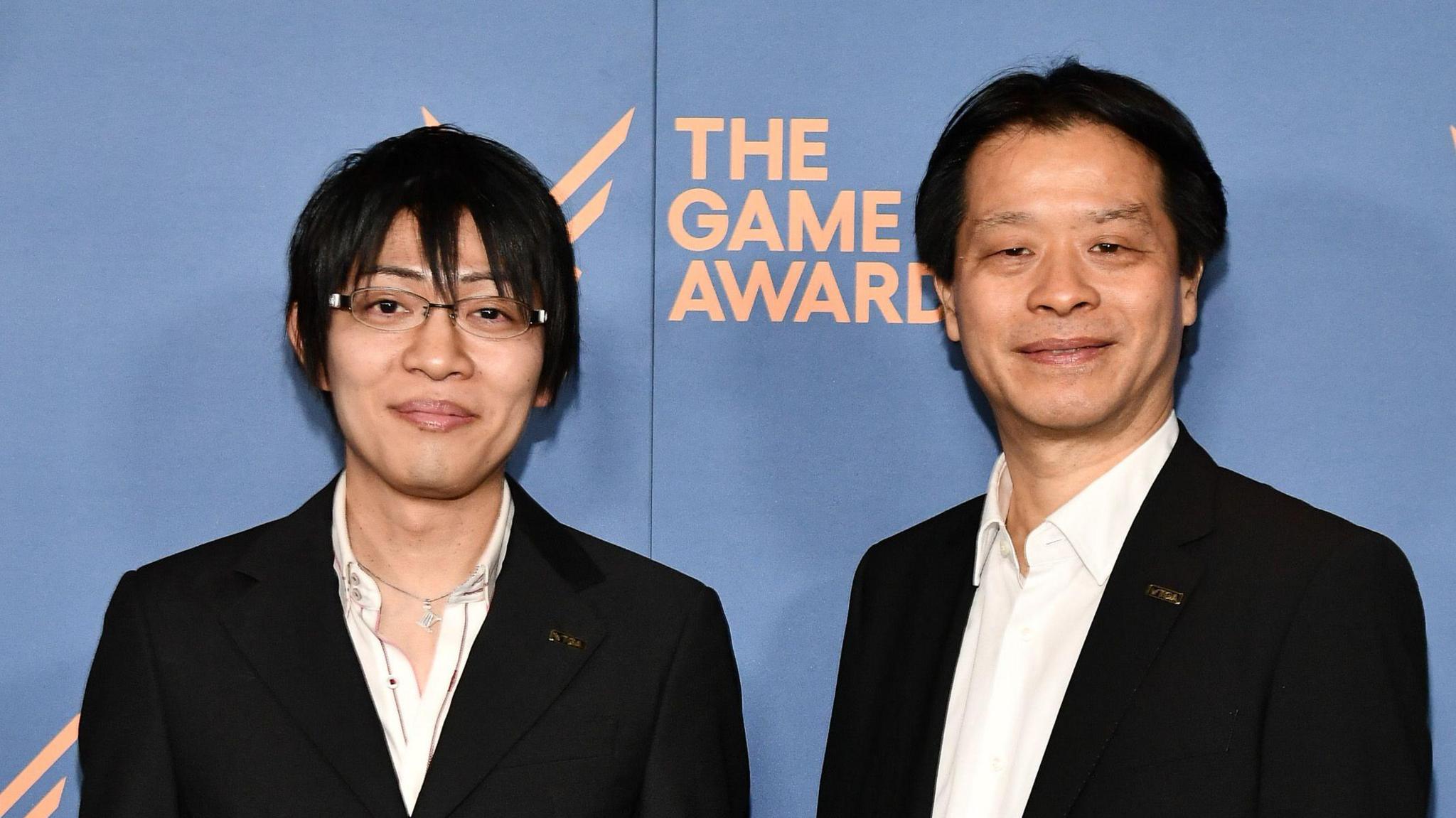 Two men in black suits and white, open-collared shirts, pose for a photo in front of a Game Awards hoarding. The younger man on the left has a mop of black hair and wears glasses, while the older man on the right has a shorter, neatly trimmed style.