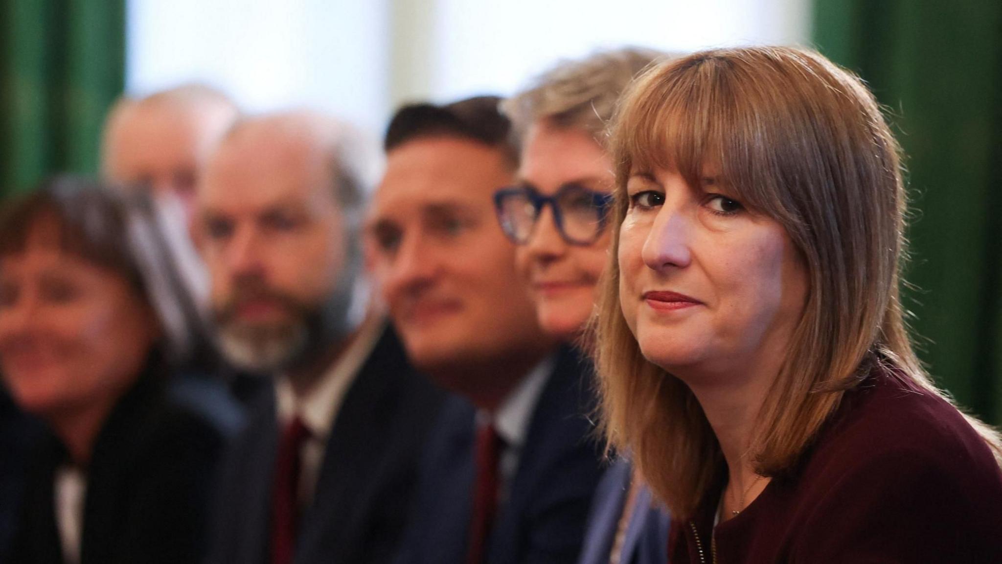 Rachel sits on the right of the frame, wearing a dark maroon top, looking seriously into the camera. Behind her sit several cabinet ministers, who appear blurry.