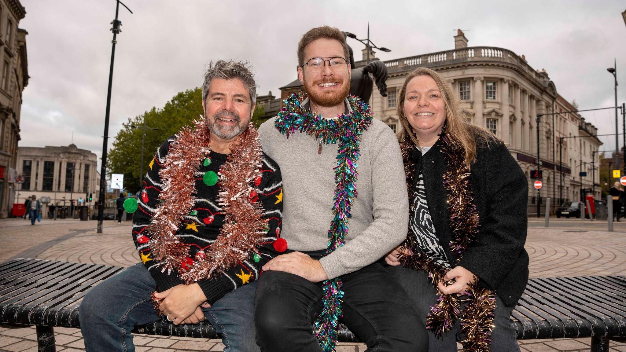 Two men and a woman are sitting outdoors on a bench. They are each adorned with tinsel and smiling for the camera. The man on the left is wearing a Christmas jumper featuring green and red knitted baubles. The man in the centre is wearing a beige jumper, while the woman on the right is wearing an animal-print top and a black jacket over it. There are large buildings behind them.