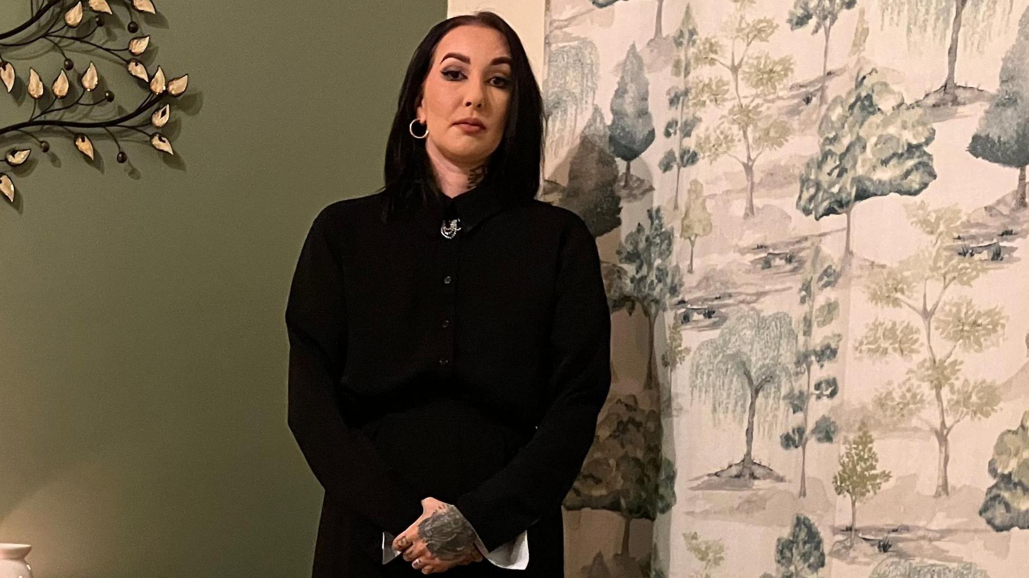 Evie Mawdsley, wearing a long black coat, stands next to an empty Moses basket. There are some lit candles on a sideboard.