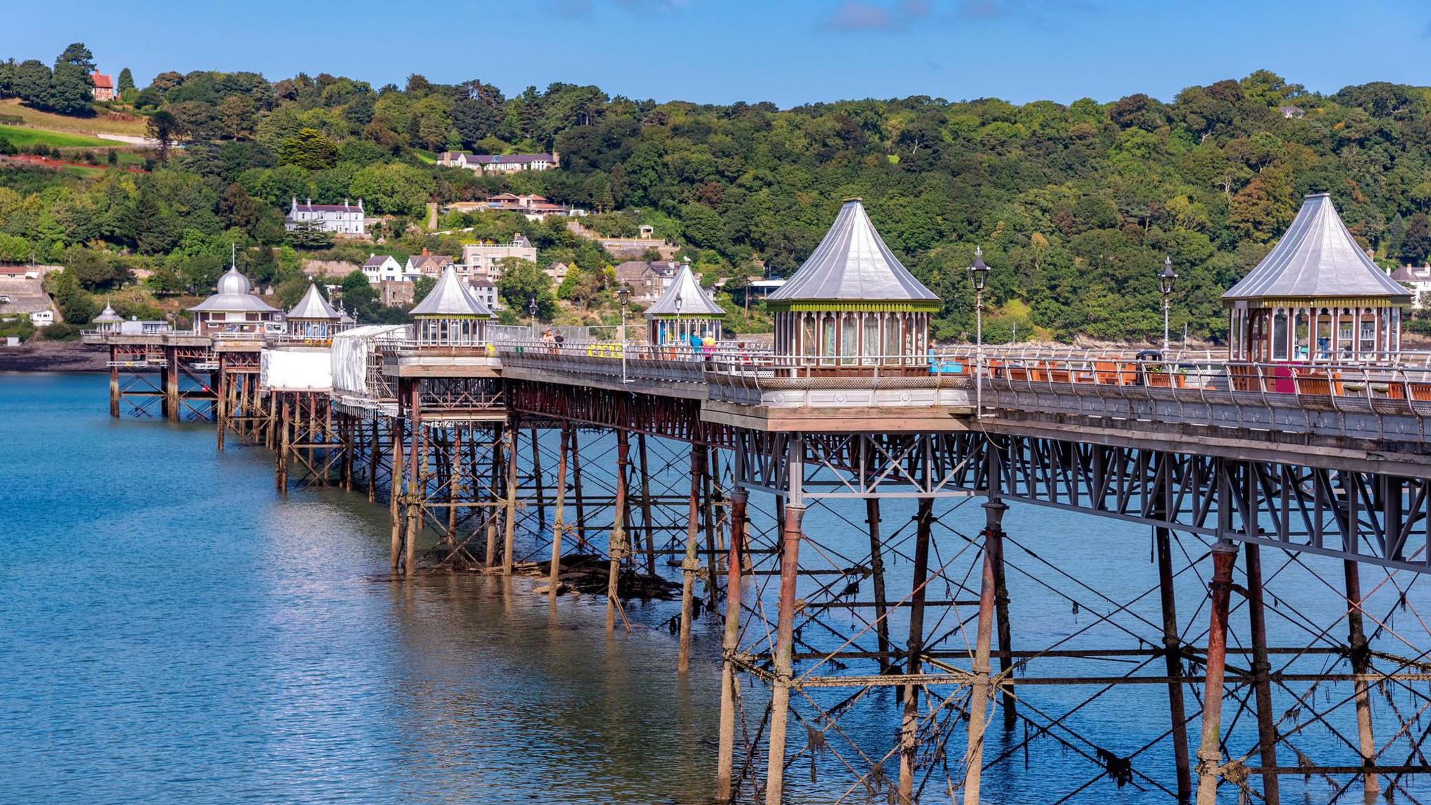 A side view of Garth Pier, with the sea below, and green hills in the background.