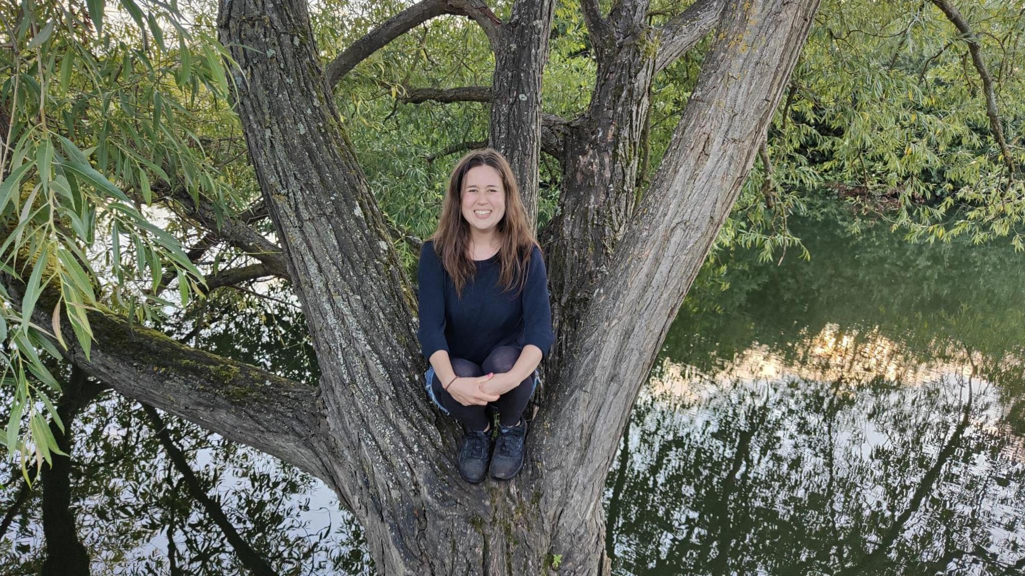 Bryony Parrish smiling for the camera while sitting in a tree. There is a body of water underneath.