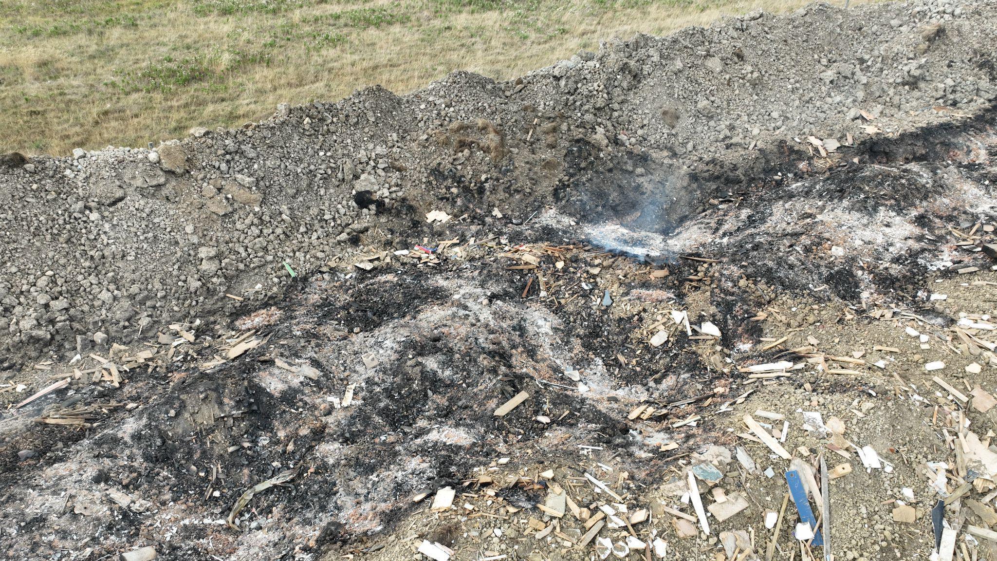 The smouldering remains of rubbish on blackened soil. There is grass in the background on the field, that has been untouched by burning.