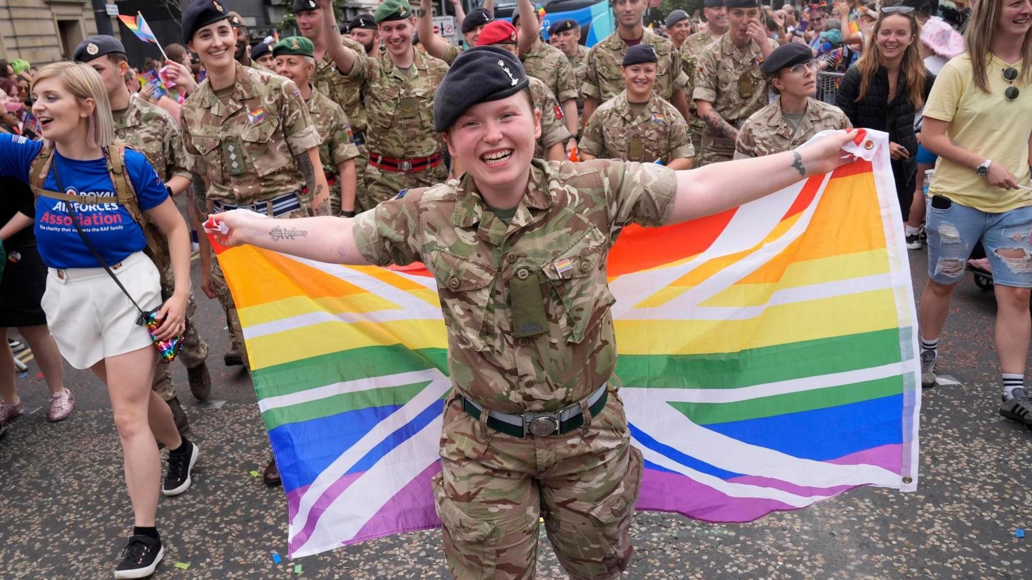 A person weather camouflage gear holds a rainbow union flag