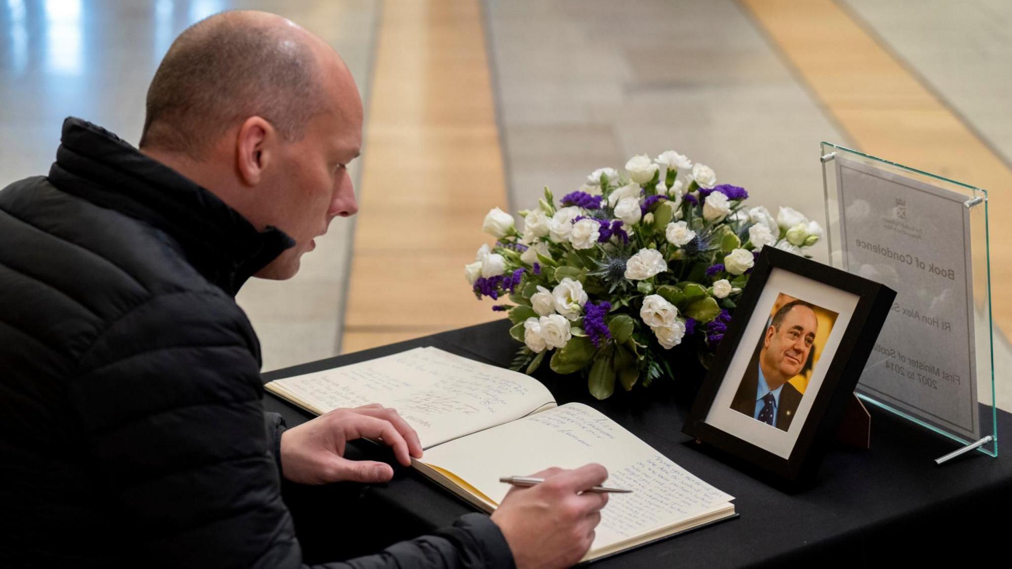 A man wearing a black jacket seated at a table writes in the book of condolence. There is a framed pictire of Alex Salmond and a bouquet of flowers on the table, which is covered in a black tablecloth