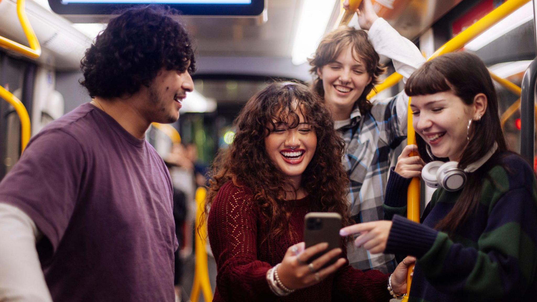 A smiling young woman surrounded by friends holds her phone on a bus