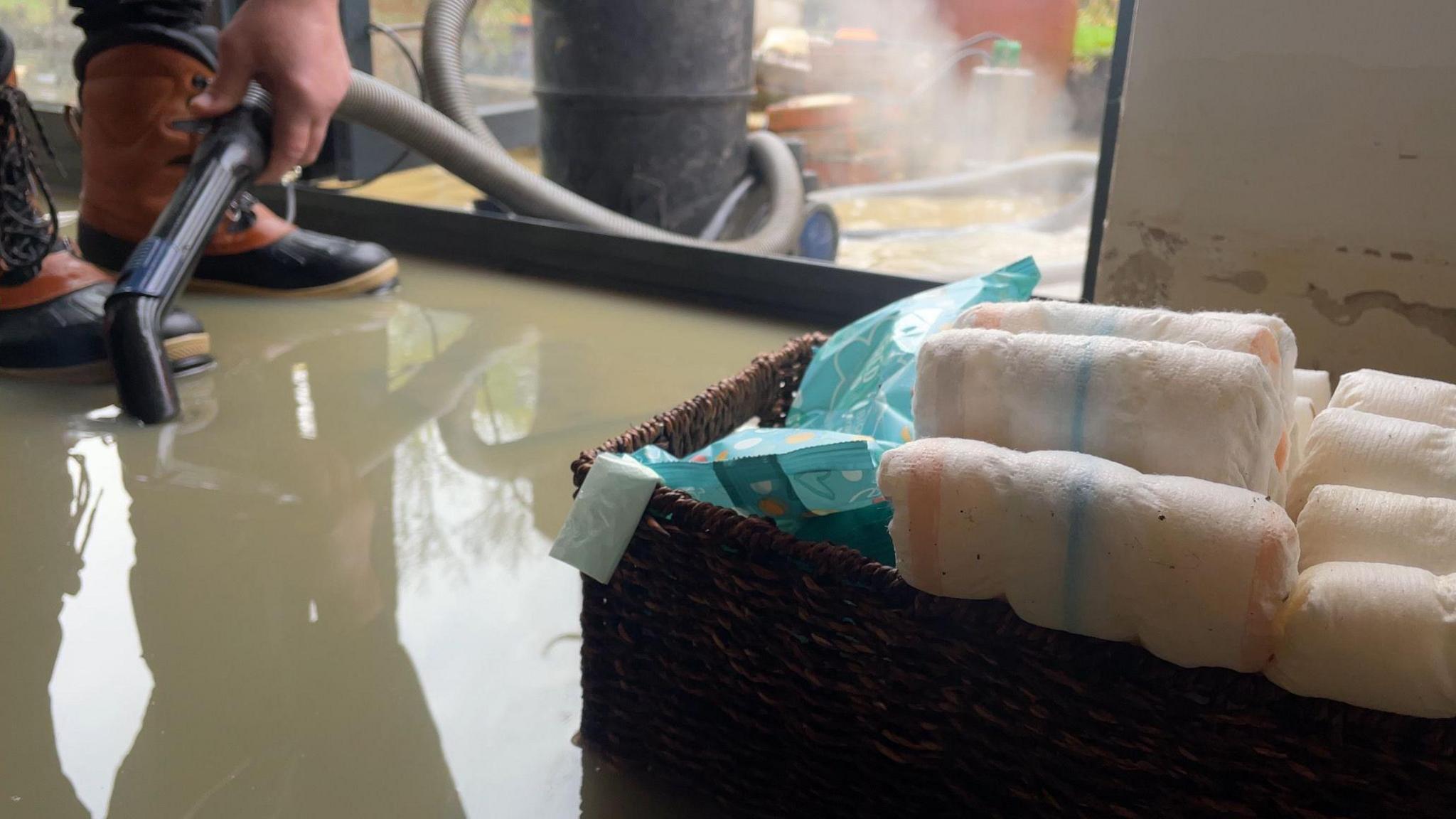 A box of nappies can be seen amid at least two inches of brown floodwater. A person wearing boots and holding a hose is pumping water from the house.