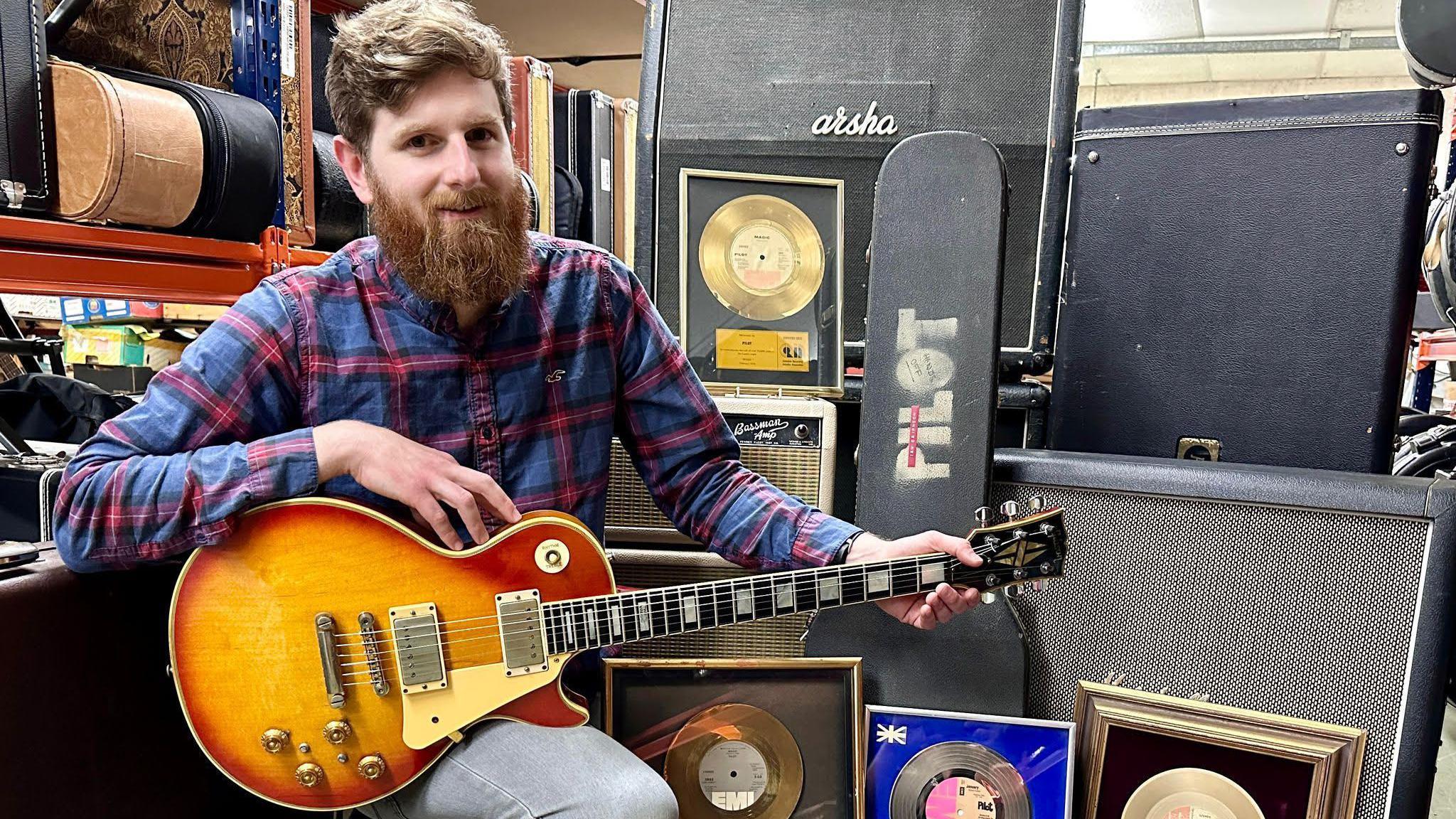 Luke Hobbs looks at the camera holding the guitar in front of other equipment