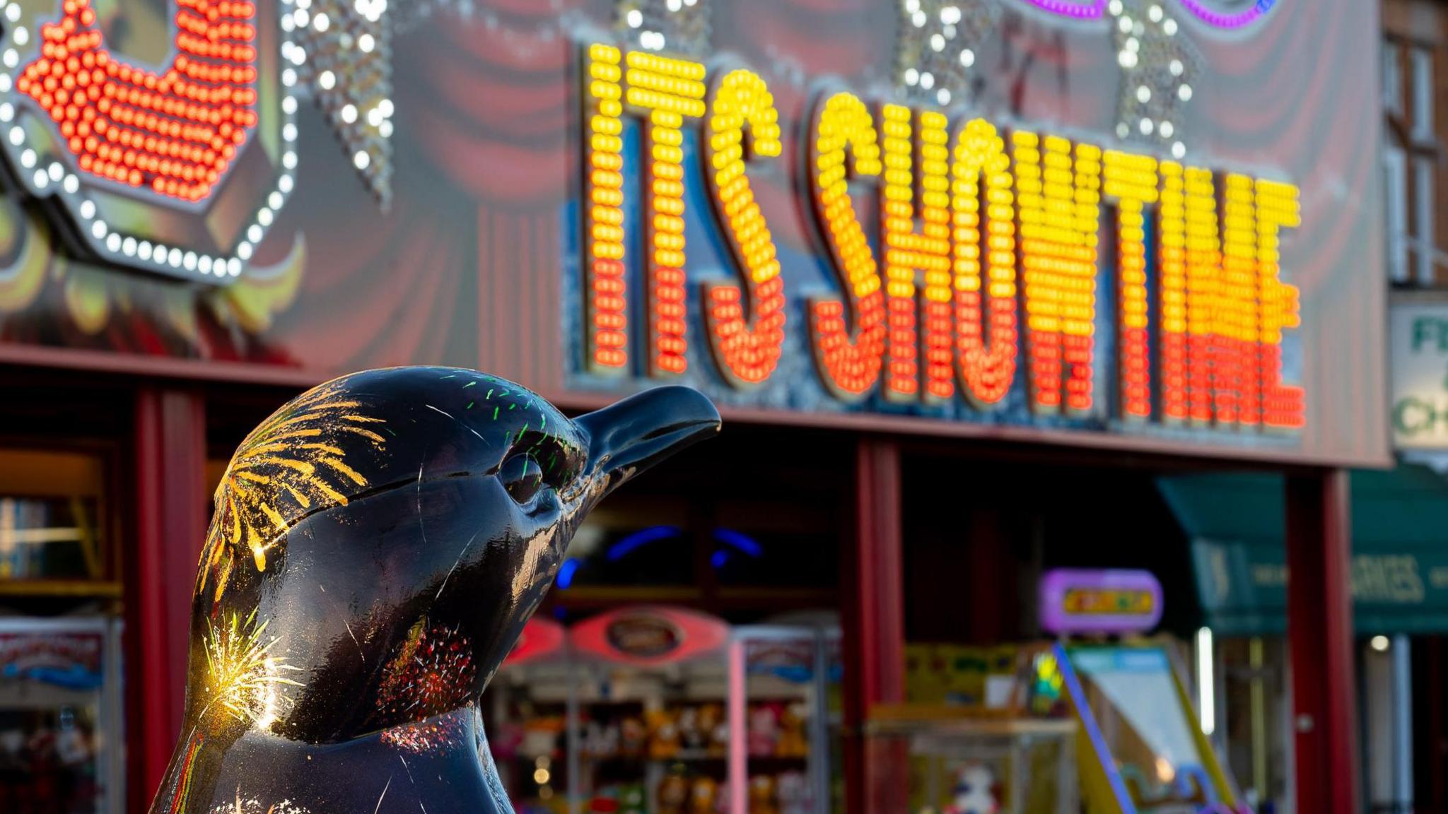 A head of a penguin sculpture, which is painted black with coloured fireworks on, is at the bottom left. In the background is the outside of an arcade which says 'Its showtime' in yellow and orange.