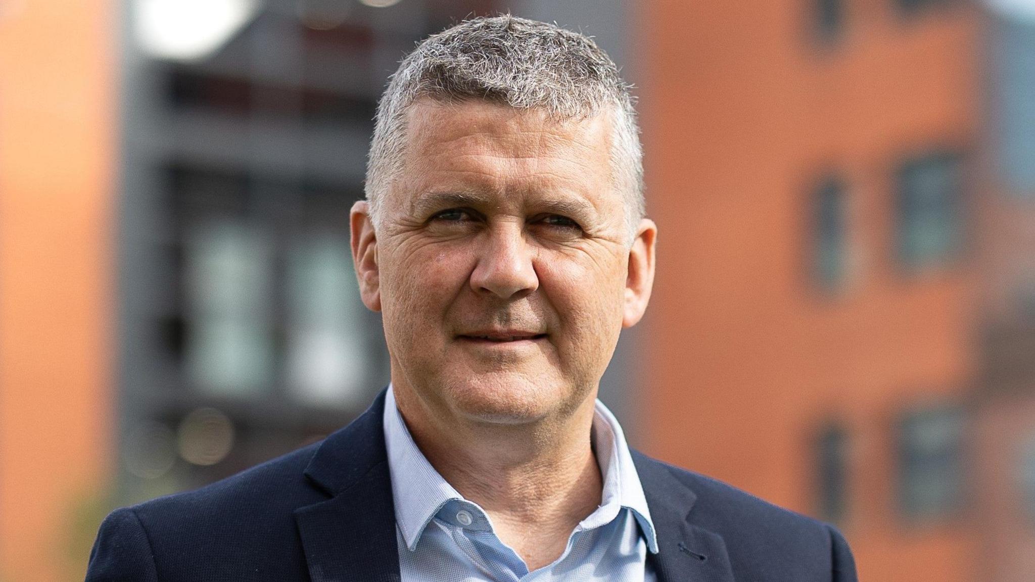 A head and shoulders picture of Mr Marsh. He is wearing a dark coloured suit jacket and a light blue shirt. He has white and black hair and is smiling at the camera.