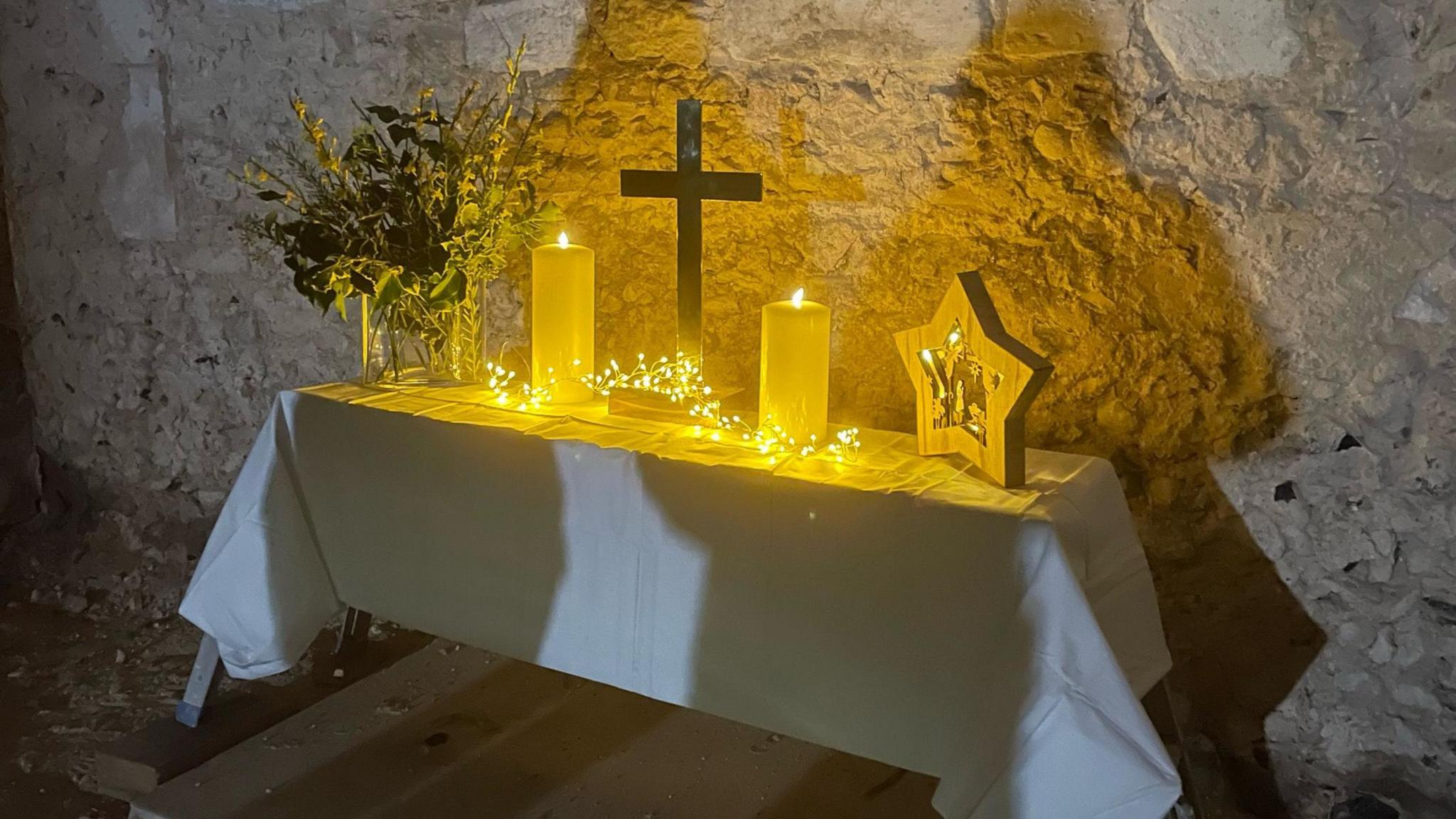 A table with a white cloth. On top of the table is two candles, fairy lights, a cross and some flowers.