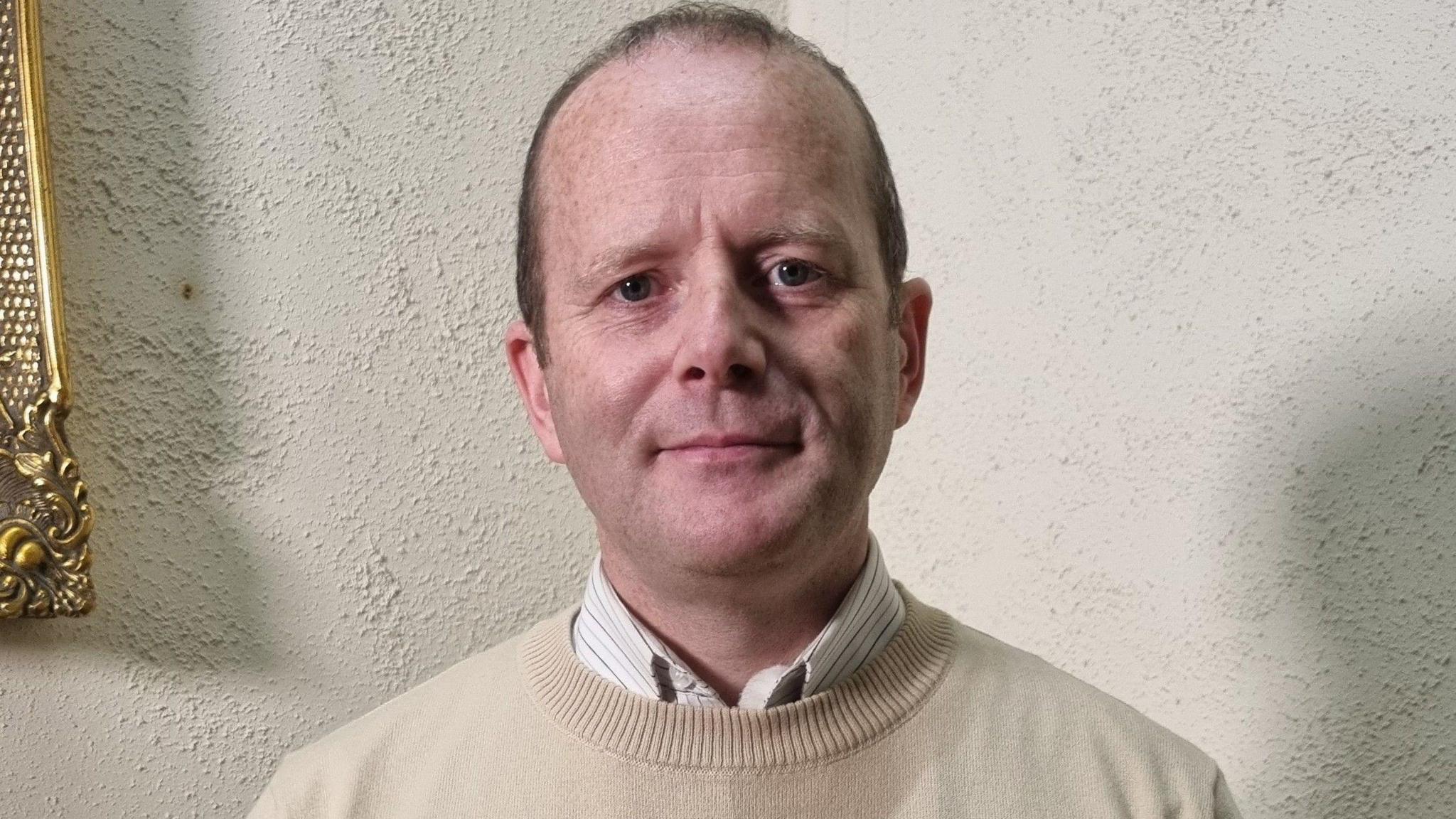 Councillor Ron McDowell smiling as he stares at the camera, he has short brown hair. He is wearing a white striped shirt and a cream jumper.