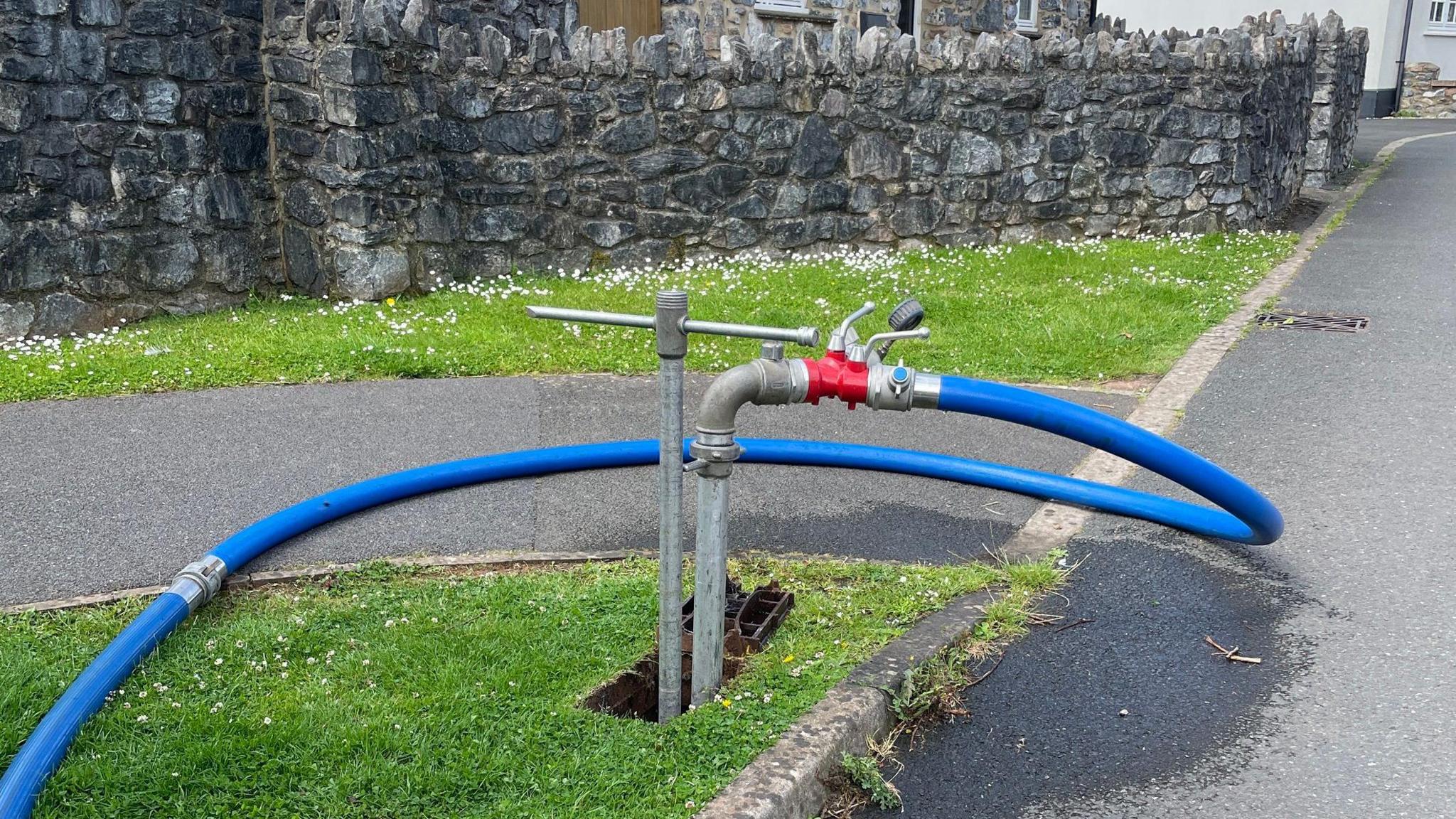 A standpipe with a tap and wide hose pipe leading from it across a footpath. Behind it is a wall and behind that is a stone house which has a conservatory.