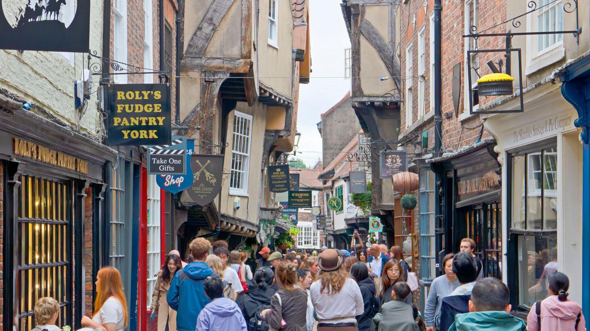 View along The Shambles in York, a narrow medieval street with timber-framed buildings housing shops and cafes. The scene shows the busy pedestrian area that retains its characteristic overhanging upper stories and historic layout.