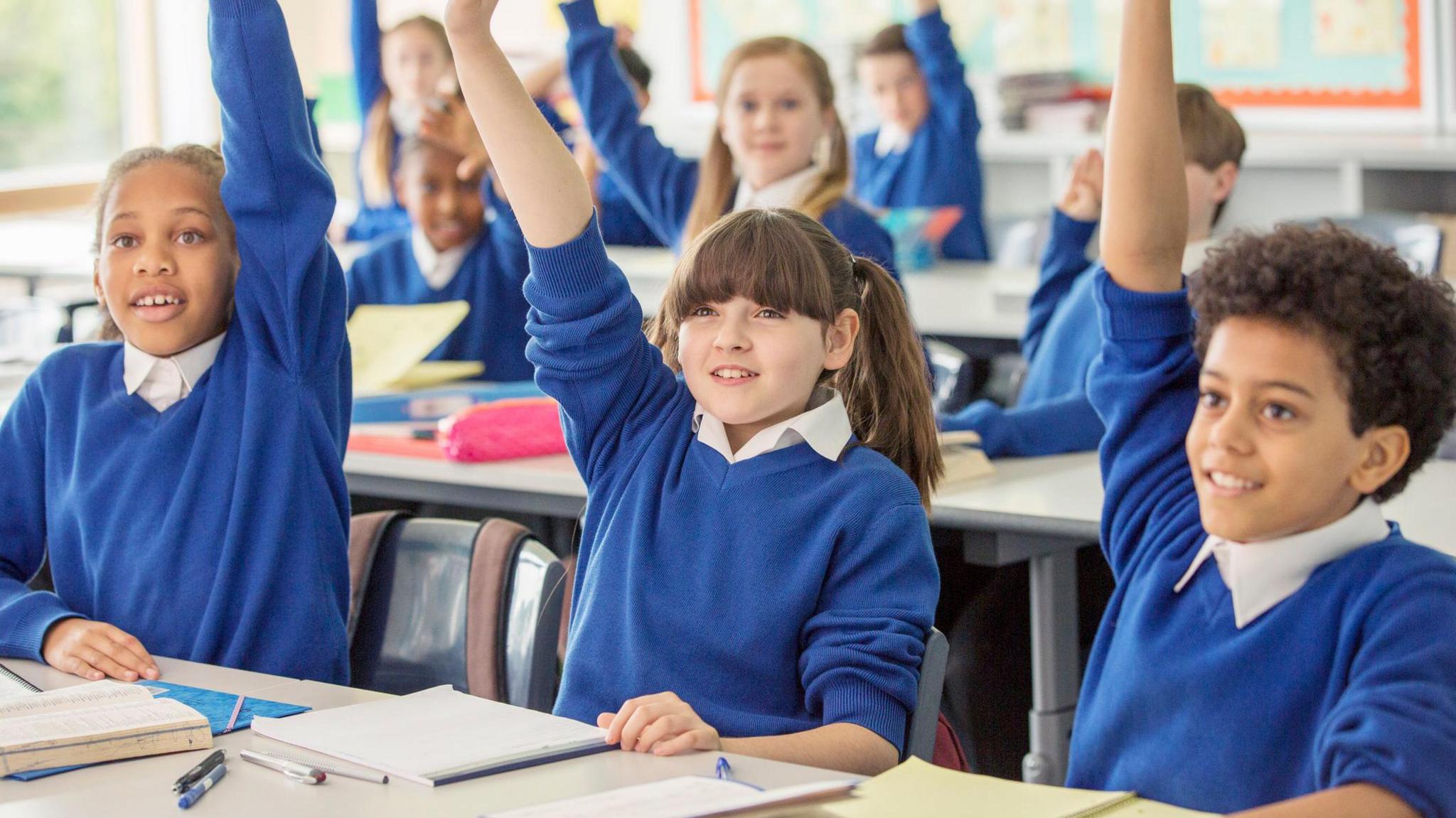 Children sit in rows at their desks in a classroom. All of them have their hands up. They are wearing white polo shirts and blue jumpers.