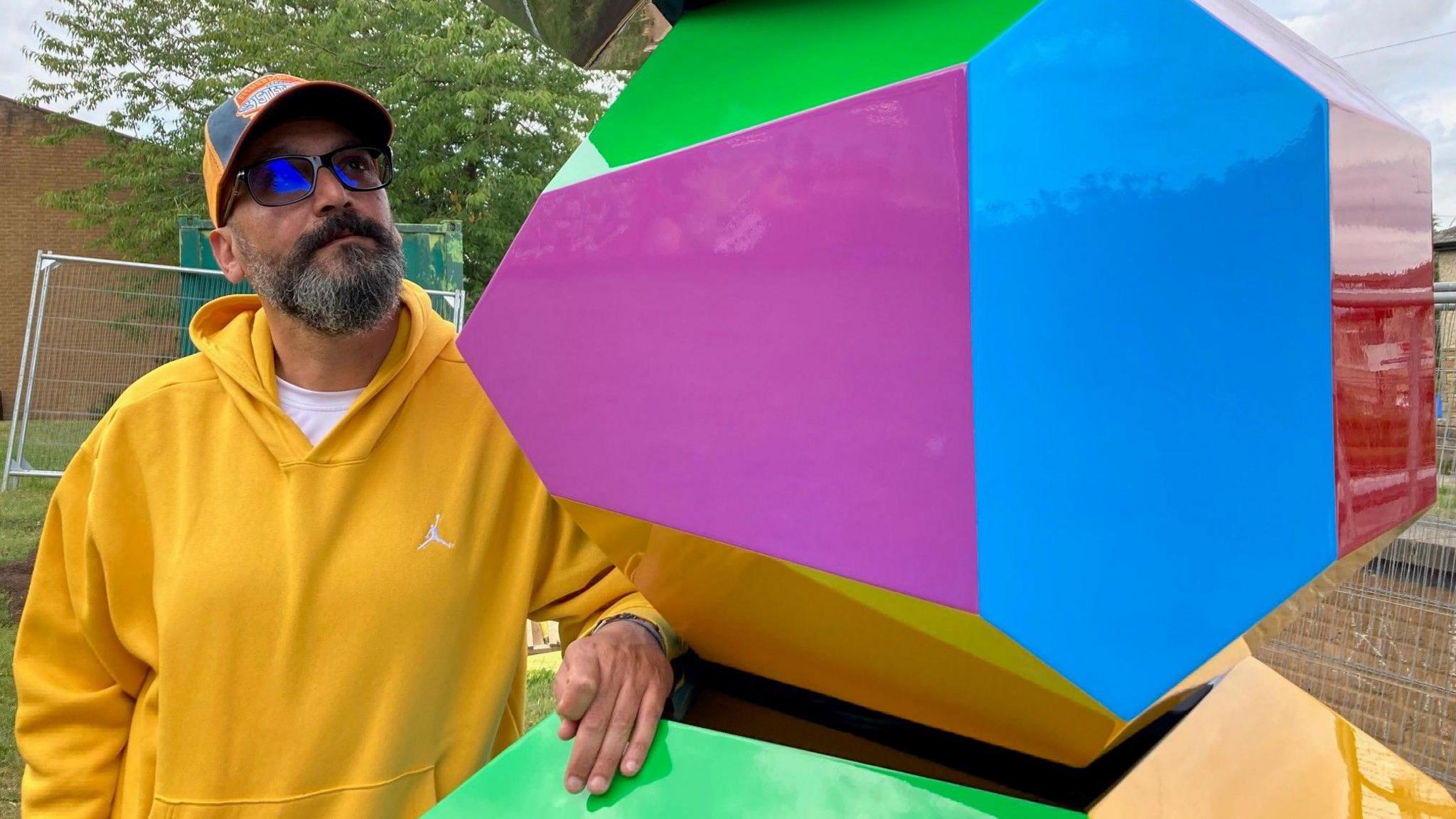 A man in a yellow hooded top and baseball cap looking at a multi-coloured block.