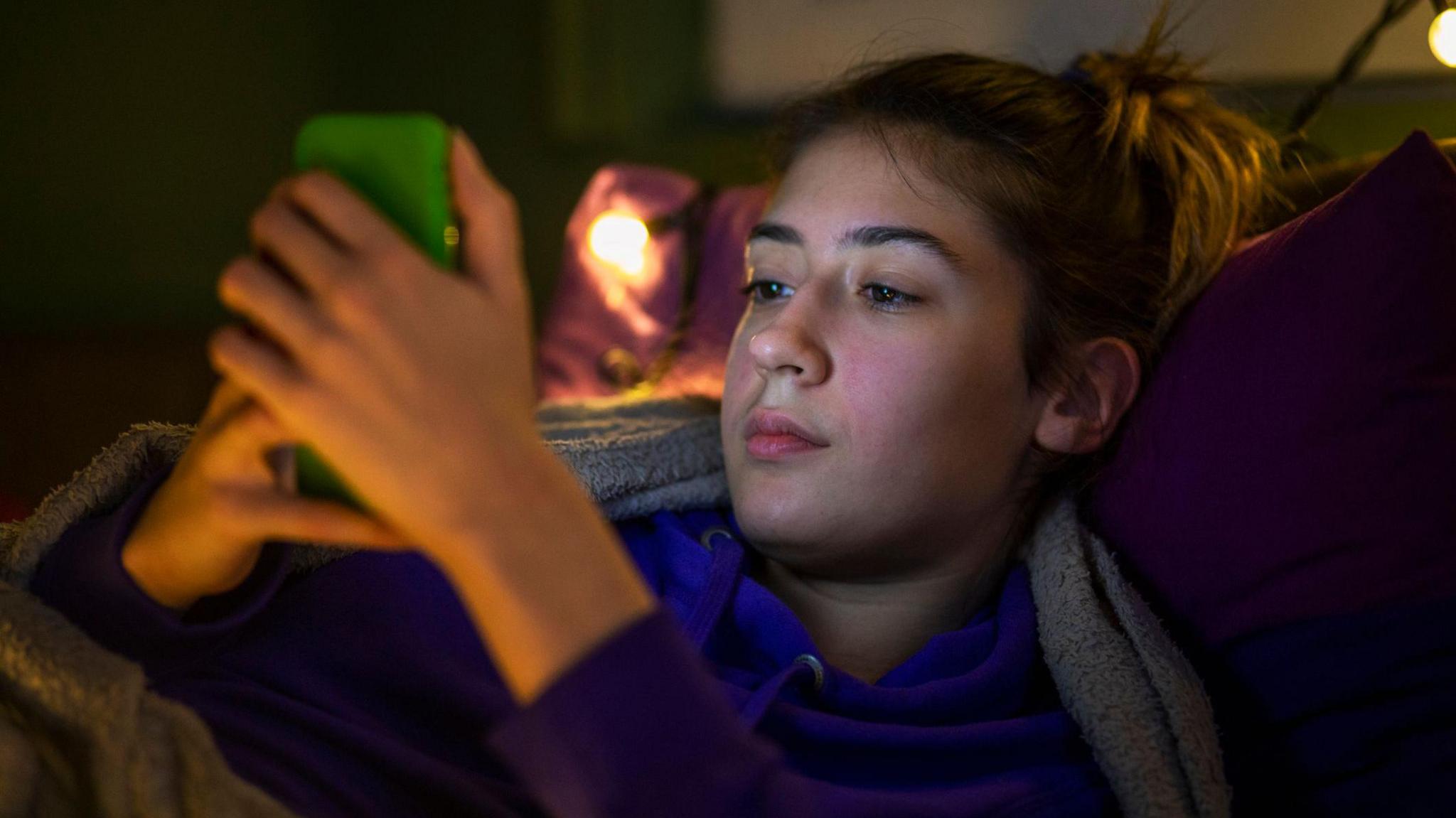 A teenage girl with brown hair tied up lies under a blanket looking at her phone. It is evening and there are fairy lights in the background. She is wearing a purple hoodie and her phone is green.