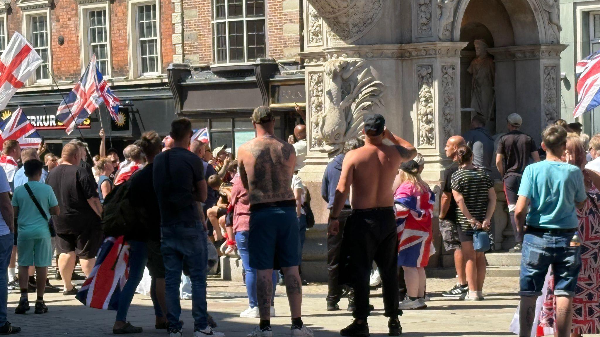 Protesters are gathered near an ornamental archway in the town. Some of the men have their tops off and are wearing baseball caps. A girl is draped in a Union flag and a woman is wearing a dress with a Union flag pattern.