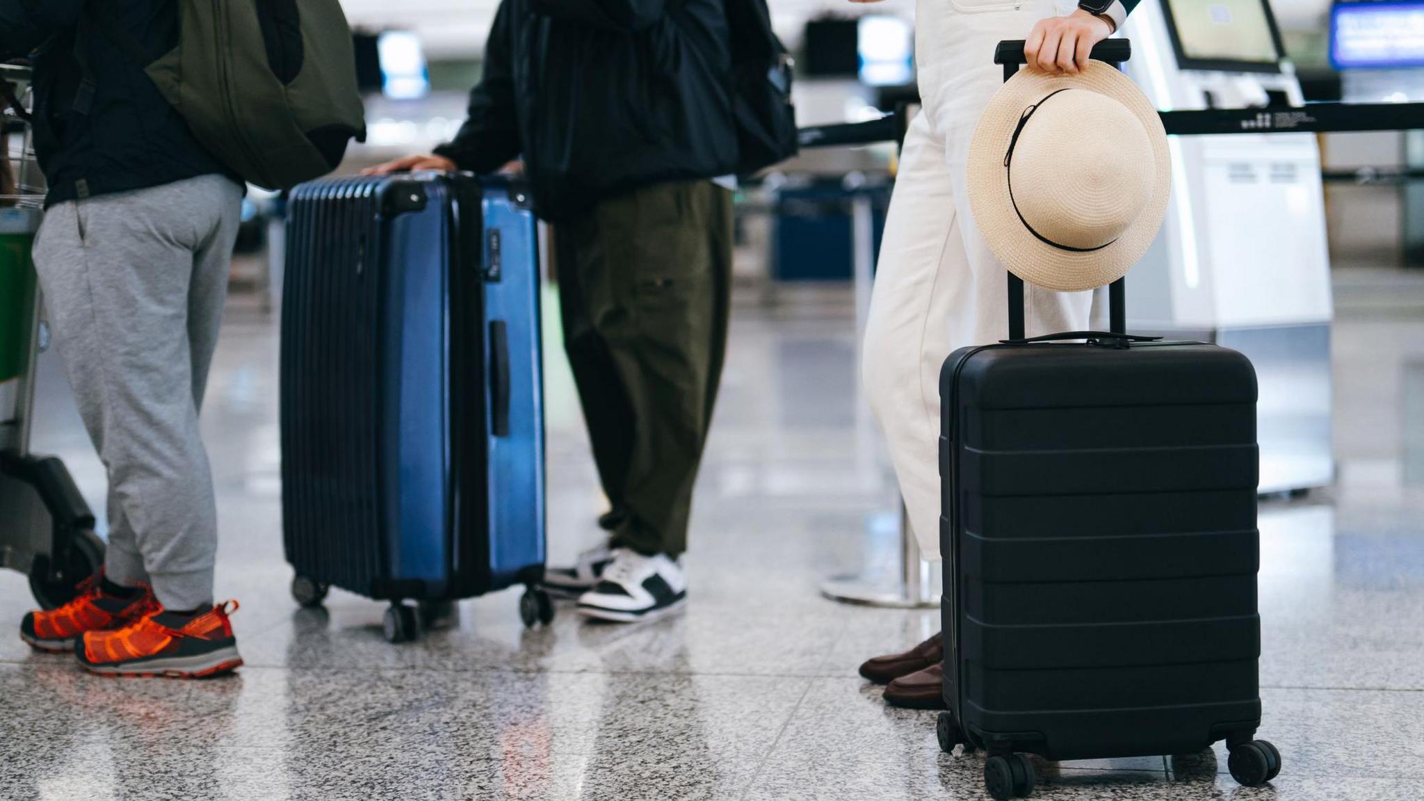 Three people wait in a line at an airport. The first person is wearing grey tracksuit bottoms, with orange and black shoes. The second person is wearing green trousers and black and white shoes with a large blue suitcase. The person behind is wearing white bottoms and holding a hat and a black suitcase.  