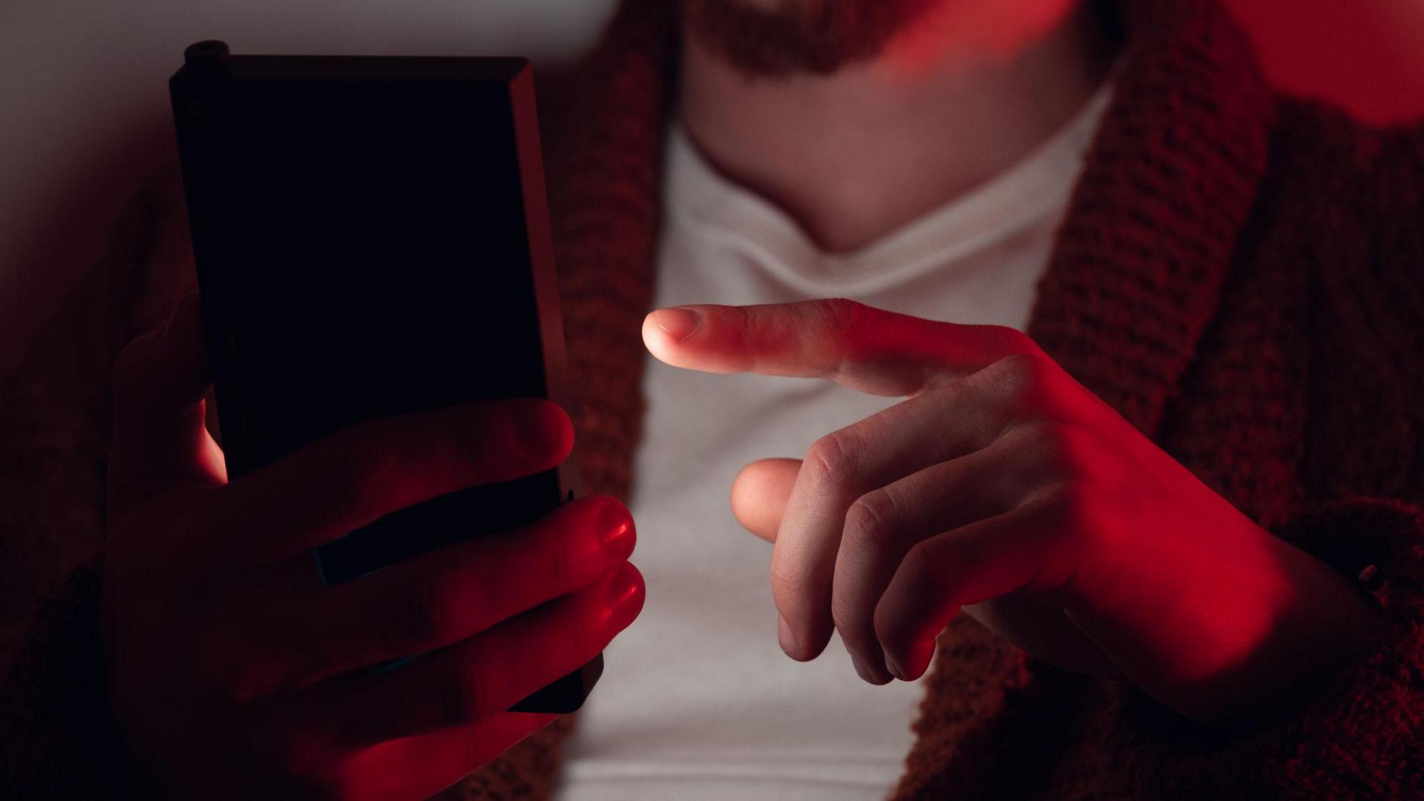 A person holding a phone and hovering a finger next to it. There is a red light on the person, whose face cannot be seen.