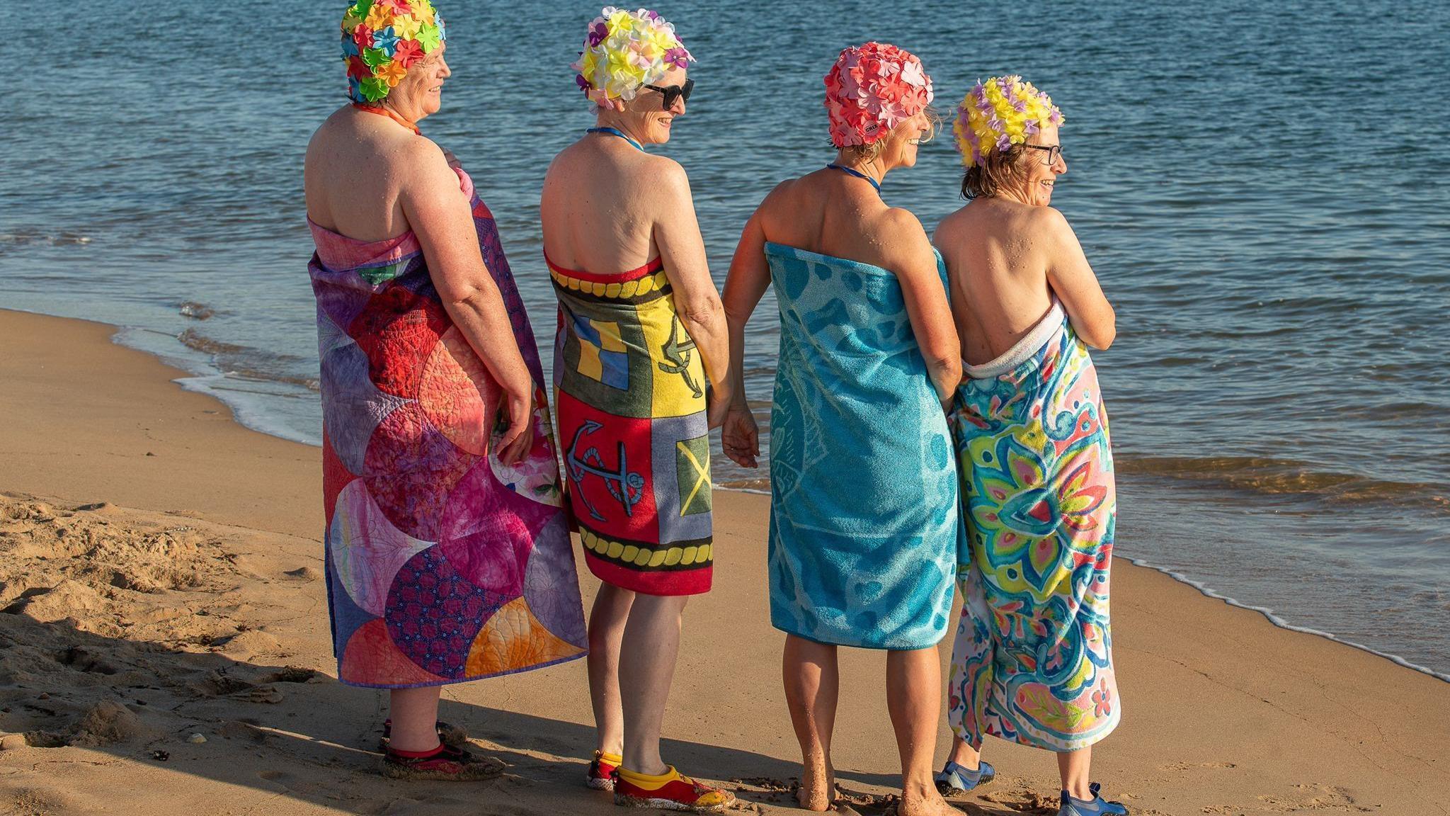 Four women wrapped in colourful towels with colourful swimming caps are standing looking out to sea and laughing.