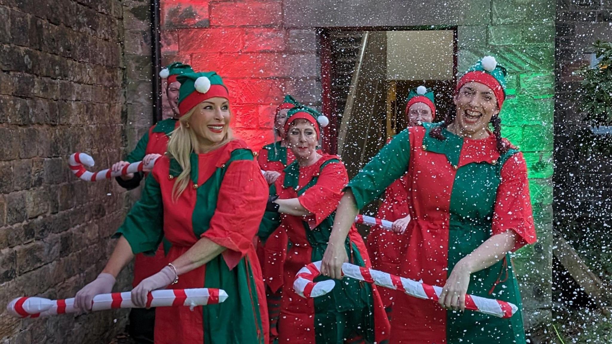 A number of women dressed as elves dancing with inflatable candy canes. The canes are white and red while the elf costumes are green and red. There is fake snow appearing from the right of the frame.