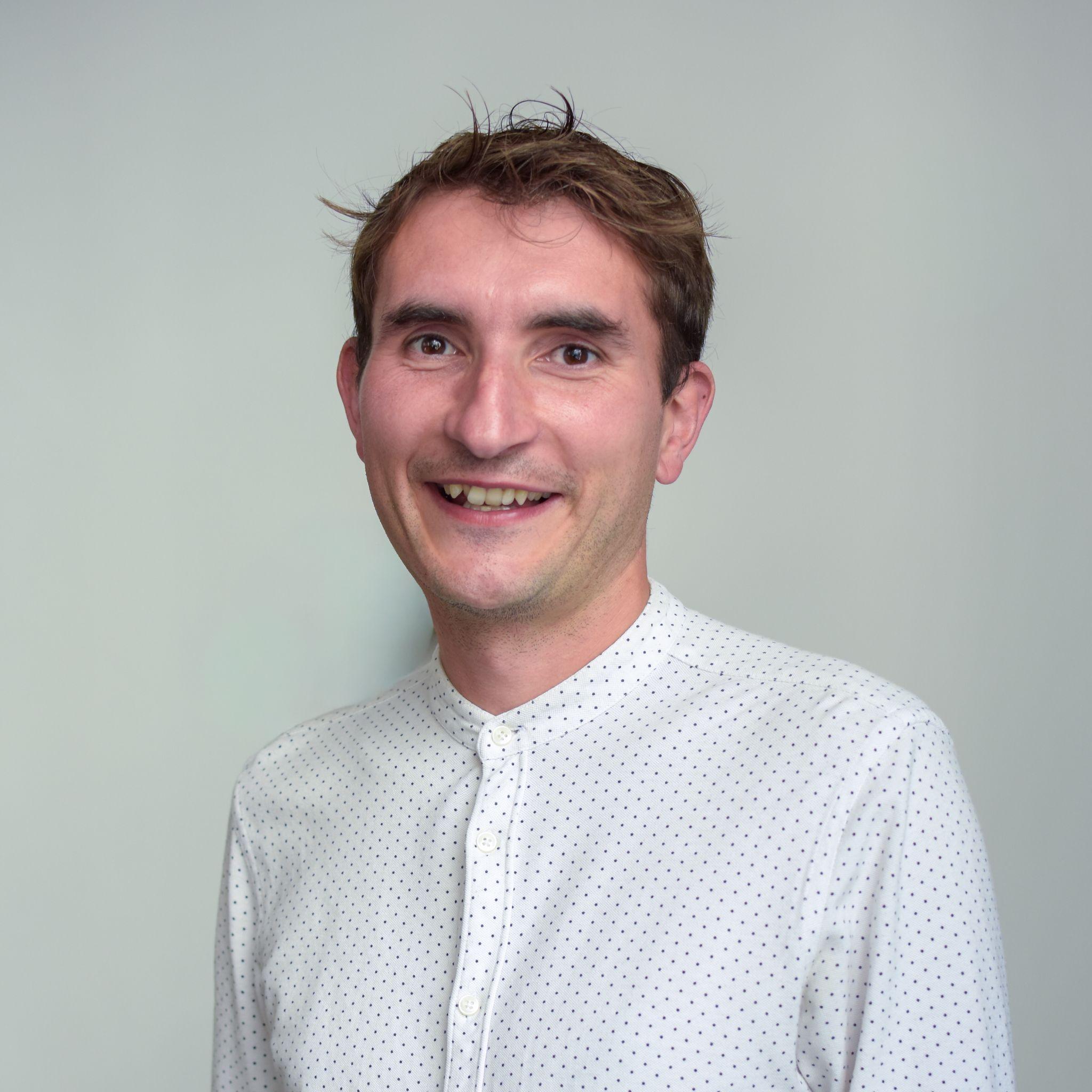 A man smiles at the camera. He is wearing a collarless white shirt buttoned up. The shirt has small dots on it. The man has brown hair and is clean-shaven.