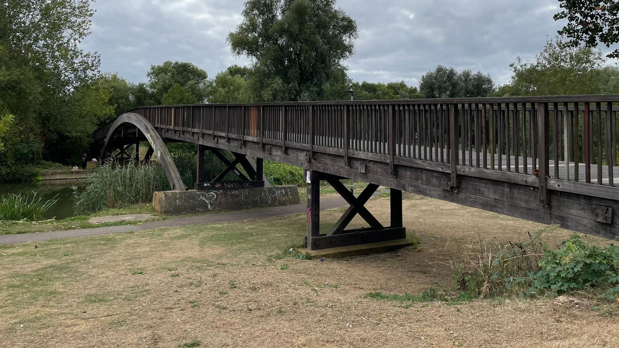 A wooden bridge stretches over a grassy area and a green river to another grass area on the other side.
