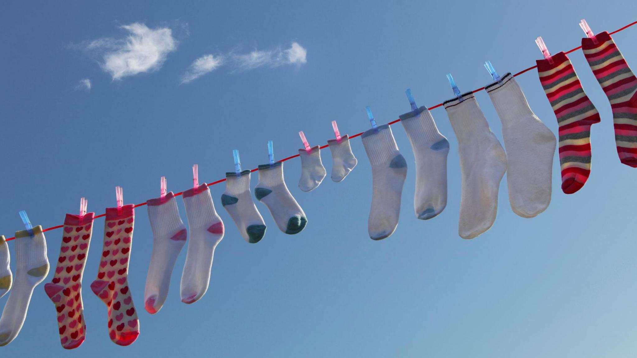 Pairs of wet socks hanging on a washing line. The washing line is red and the socks are being hung up with pink and blue pegs. Most of the socks are white with either yellow, pink. green or blue feet and ankles. One pair has pink and red hearts and one pair has green, pink and red stripes.