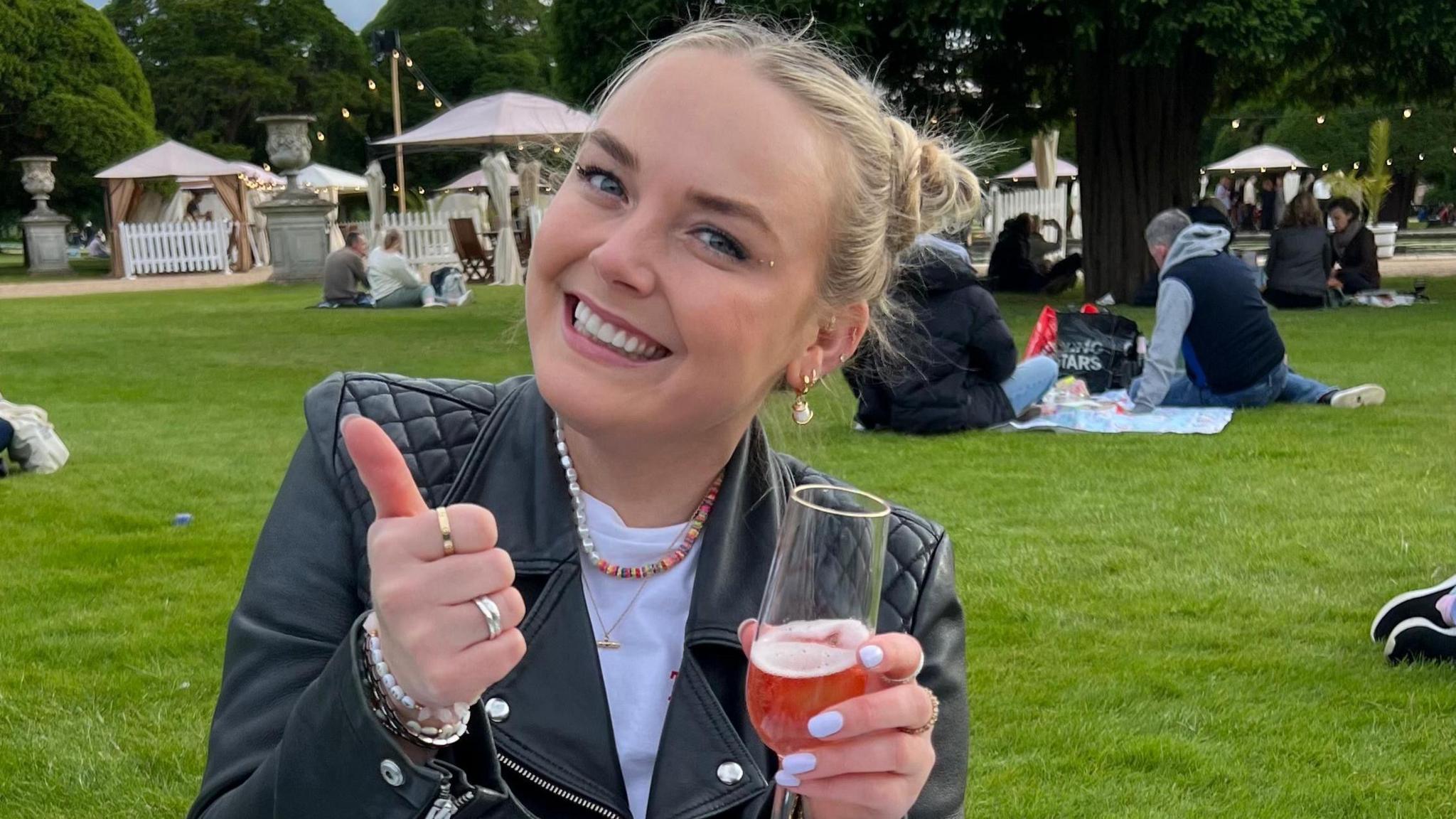 Georgie does a thumbs up to the camera, wearing a leather jacket, and holding a drink.