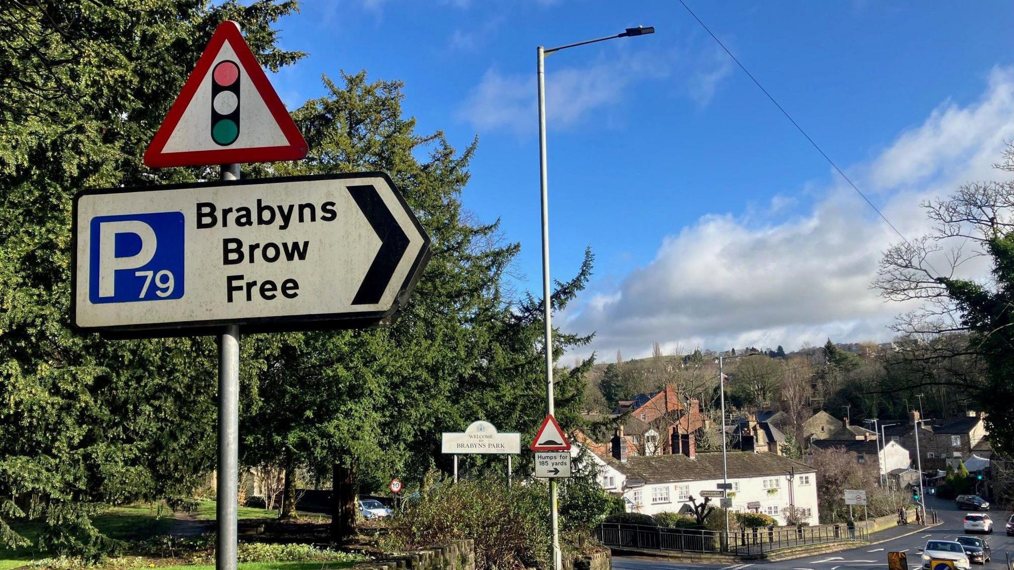 A traffic light road sign above another sign which reads 'Brabyns Brow Free' on it, can be seen in the foreground, with trees and a hilly road with cars driving on it in the background on a sunny day with blue skies. 

