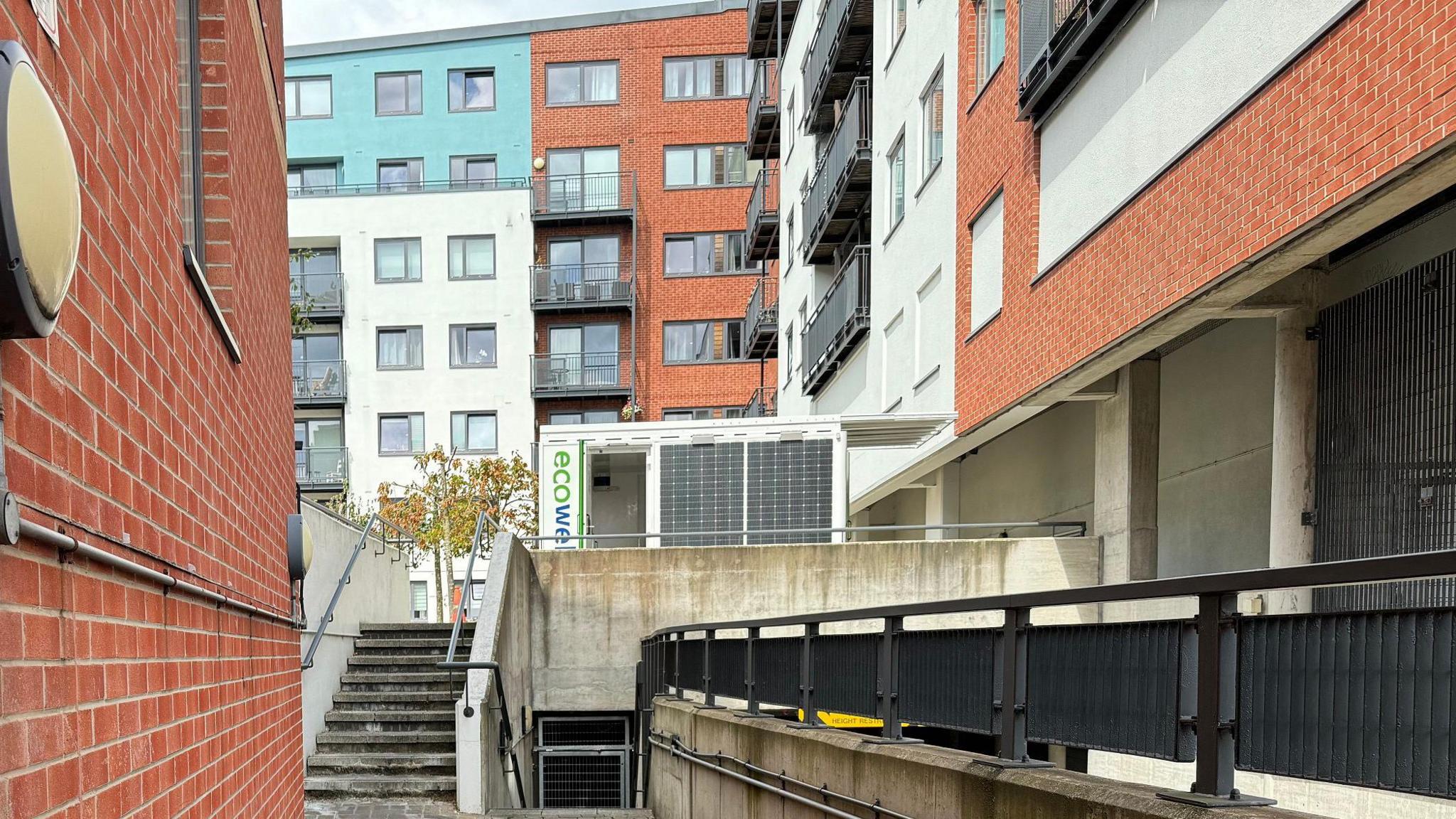 The Courtyard in Camberley. There are several blocks of flats which are red brick and also blue and white in colour. Some have balconies. Grey concrete steps lead up to a white hut, which is where the 'waking watch' is based.