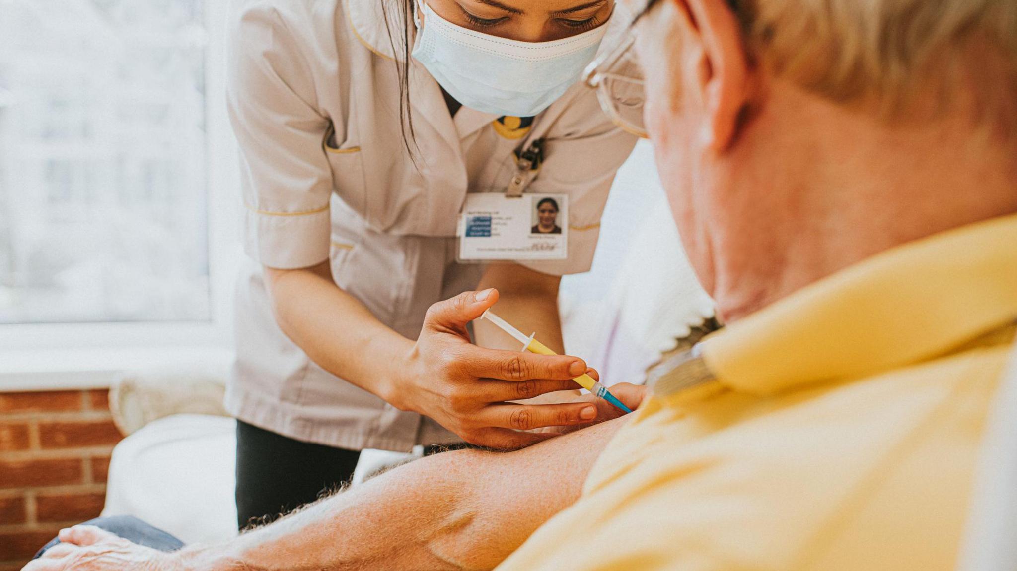 Female nurse gives an elderly male an injection in his upper arm. She wears a mask and he is wearing a yellow shirt. 