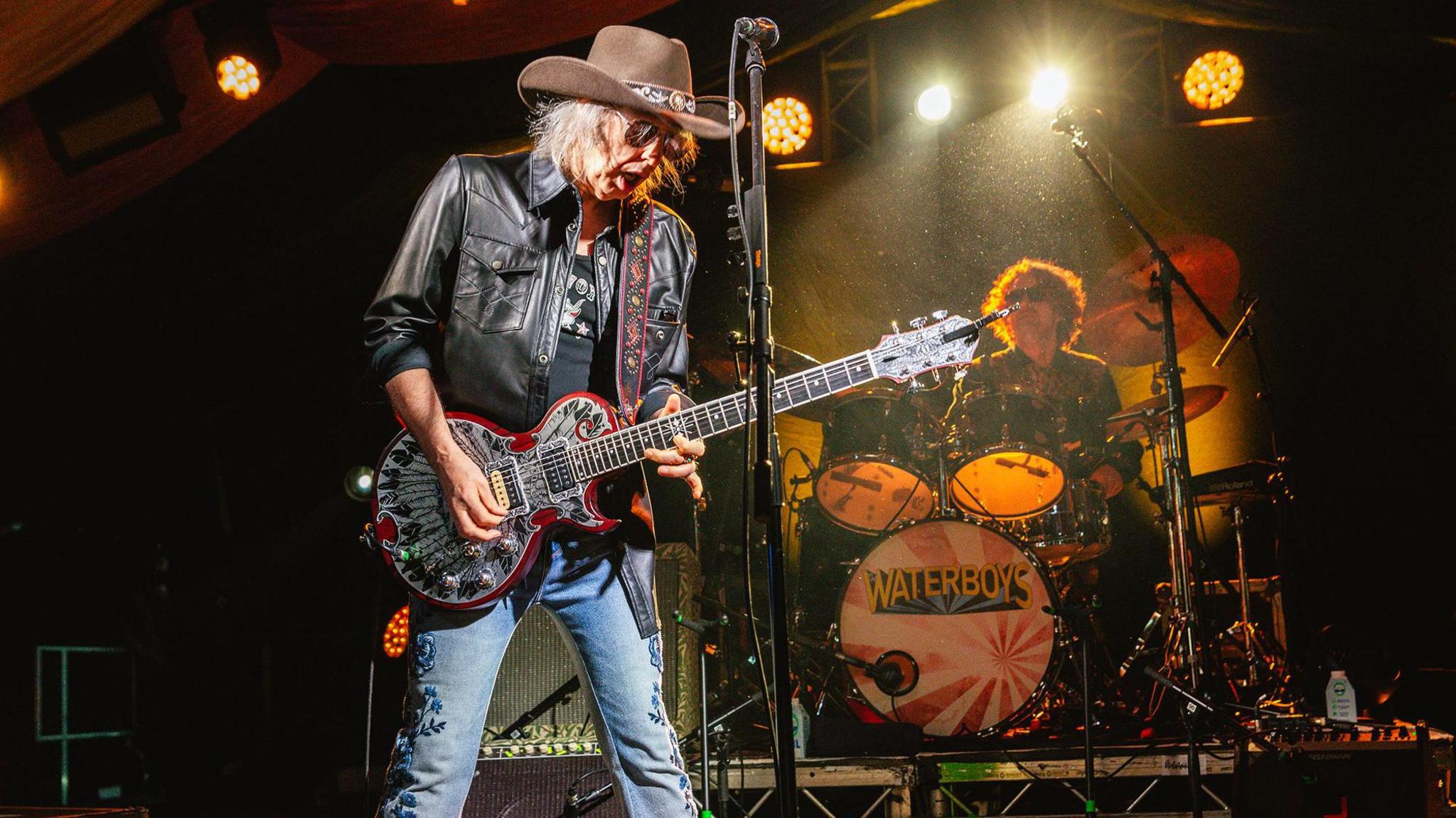 Mike Scott, lead singer of The Waterboys, wears a wide-brimmed hat, leather jacket and jeans. He is wearing dark glasses and playing a guitar. 