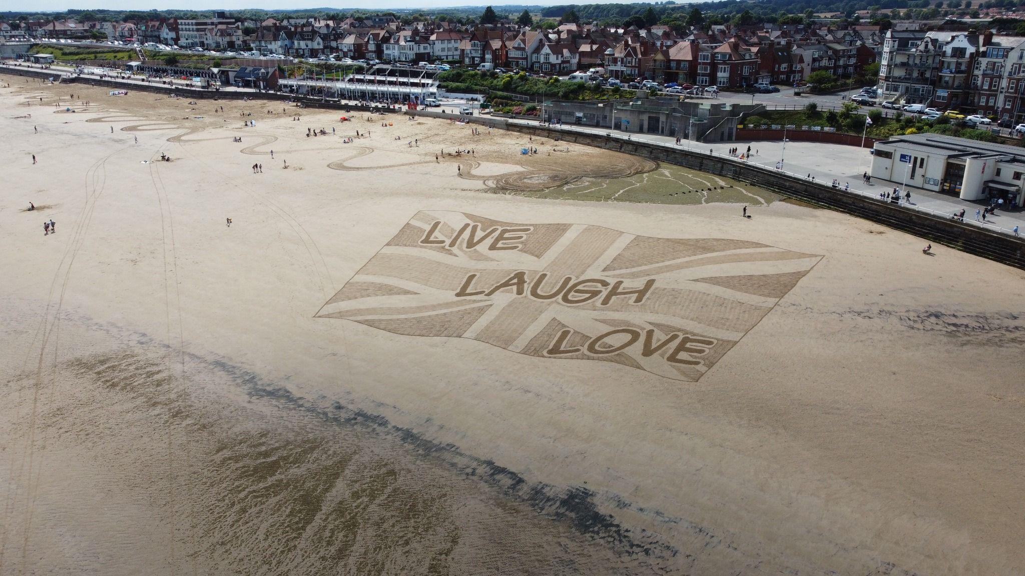 A large sand artwork on Bridlington beach, with a Union Jack flag and the slogan live, laugh, love, in a comic sans font. In the background are houses and hotels on Bridlington sea front.