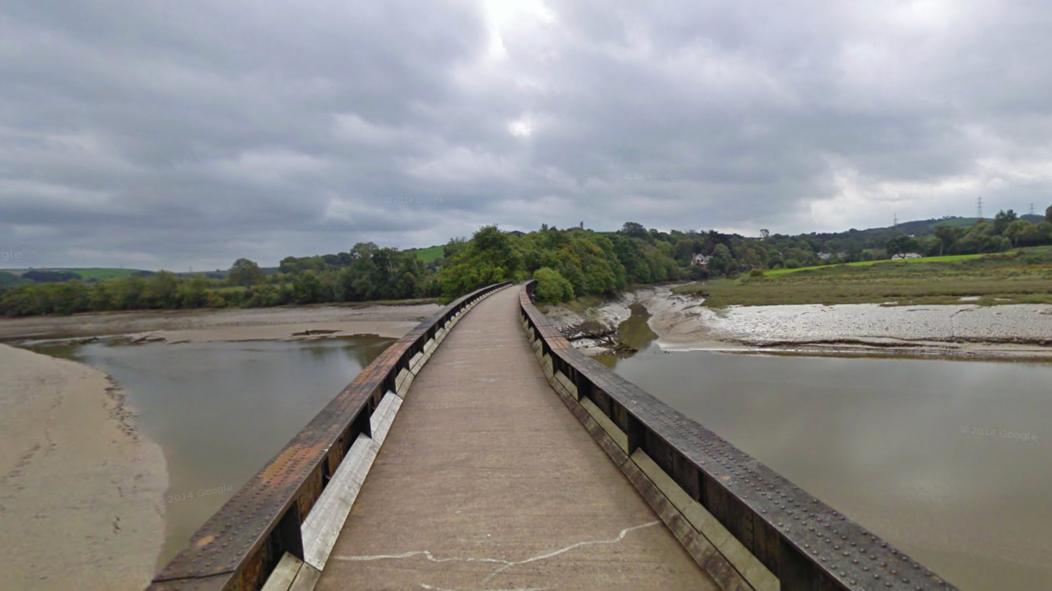 A narrow pedestrian bridge with wooden railings spans a muddy river or estuary. The sky is overcast with grey clouds, and trees and greenery line both sides of the water. No people are present in the scene.