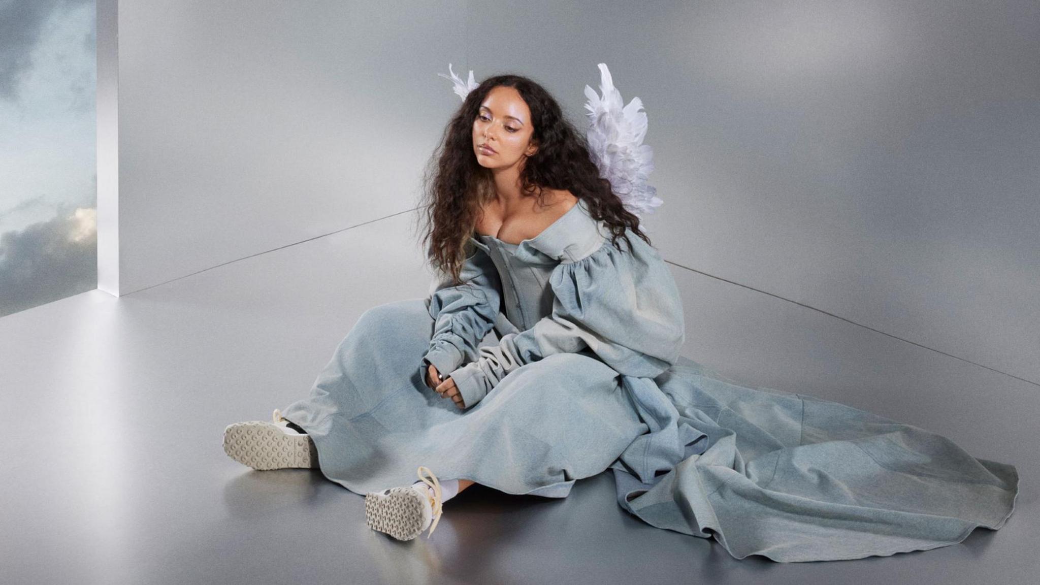 Jade Thirlwall in album artwork for her new single. Jade has long curly brown hair worns loose and sits on the floor, her hands in her lap, while wearing a washed denim gown, white trainers and angel wings 