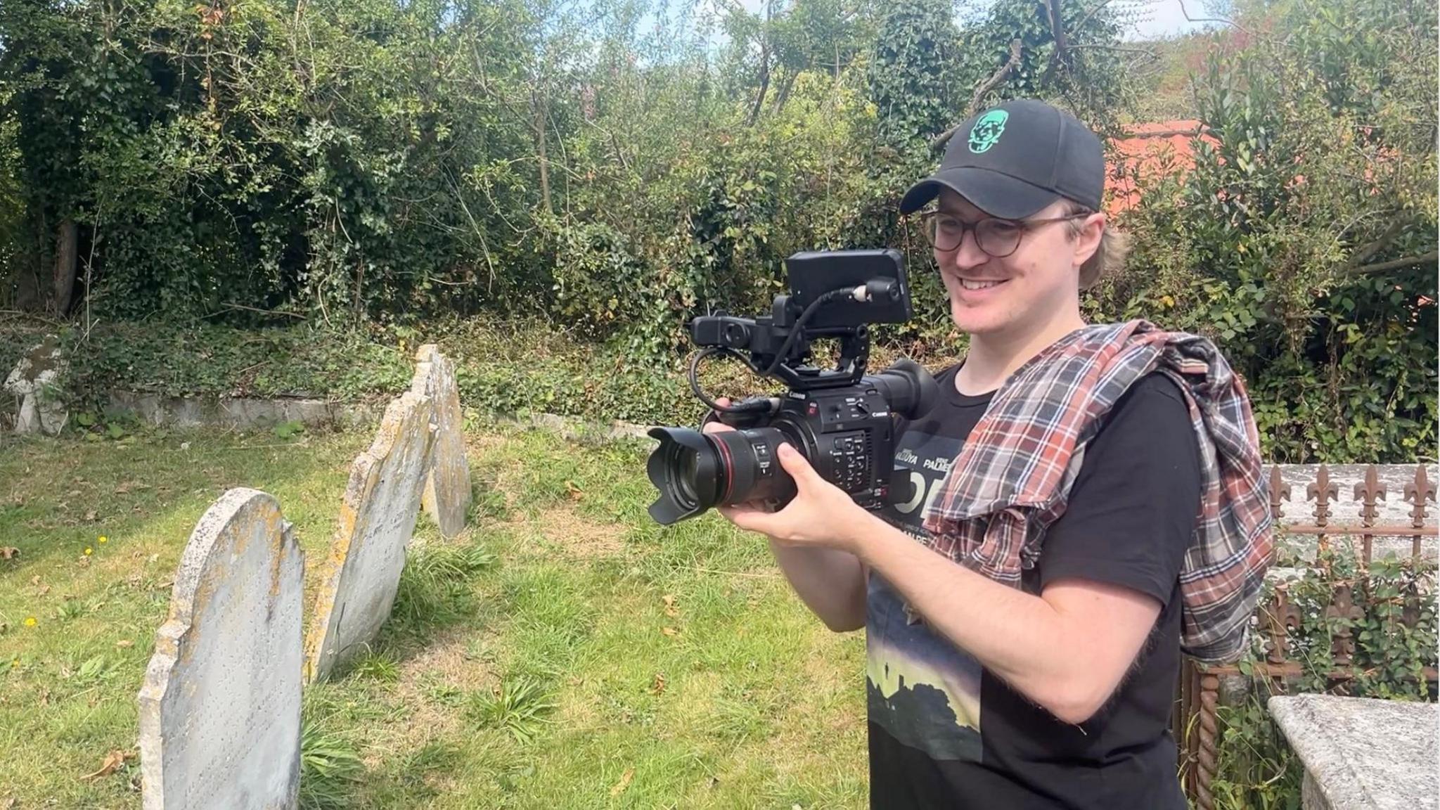 A smiling man in a black cap and T-shirt with a video camera in hand and a checked shirt draped over his shoulder. He stands in a graveyard, filming.