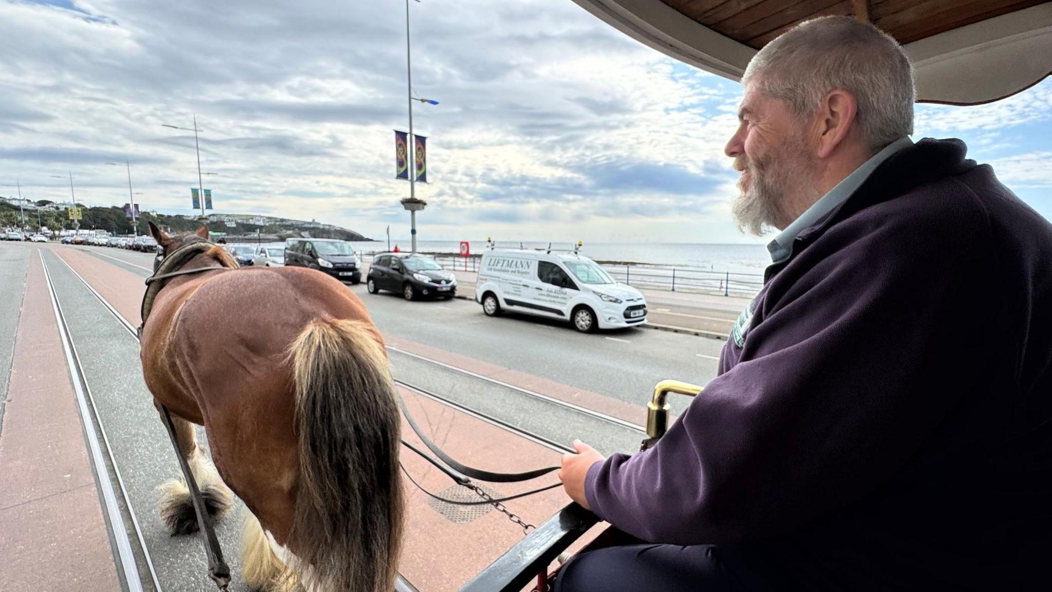 Peter Cannon on the left is at the front of the tram holding the reins of the brown horse on the left walking along Douglas Promenade with cars in the background.