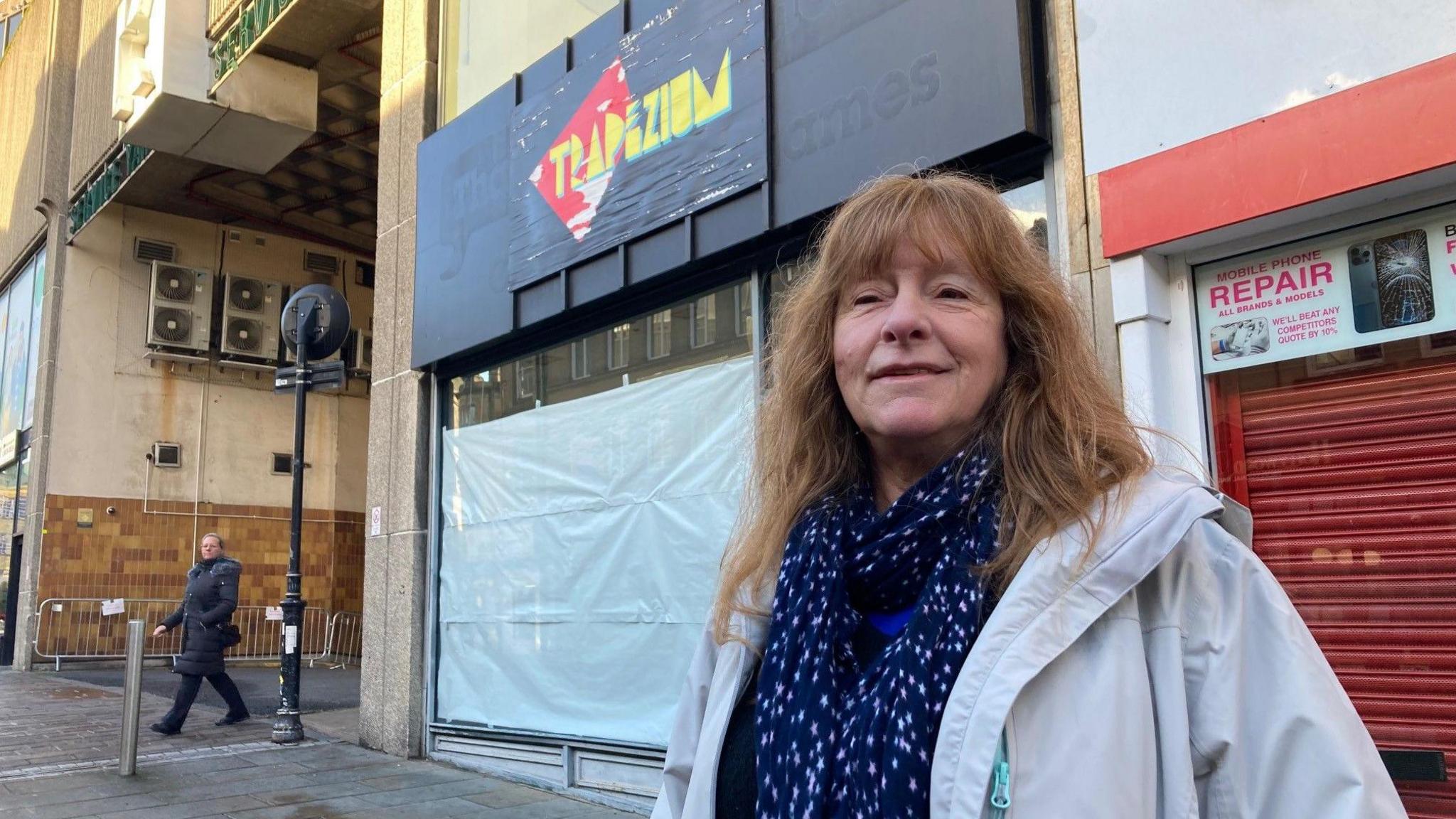 A woman with long, brown hair wearing a light grey hooded jacket and blue and white blouse standing outside a shop front which has white paper taped up inside the windows.  