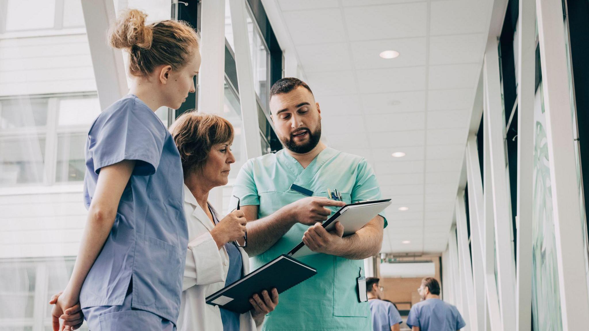 A male healthcare worker with a dark beard and green scrubs points to a chart as he talks to two female health care workers. One, an older doctor, wears a white coat, above scrubs and holds a pair of glasses in her hand. The other, a younger woman with curly light hair tied up wears scrubs and stands with her hands behind her back.