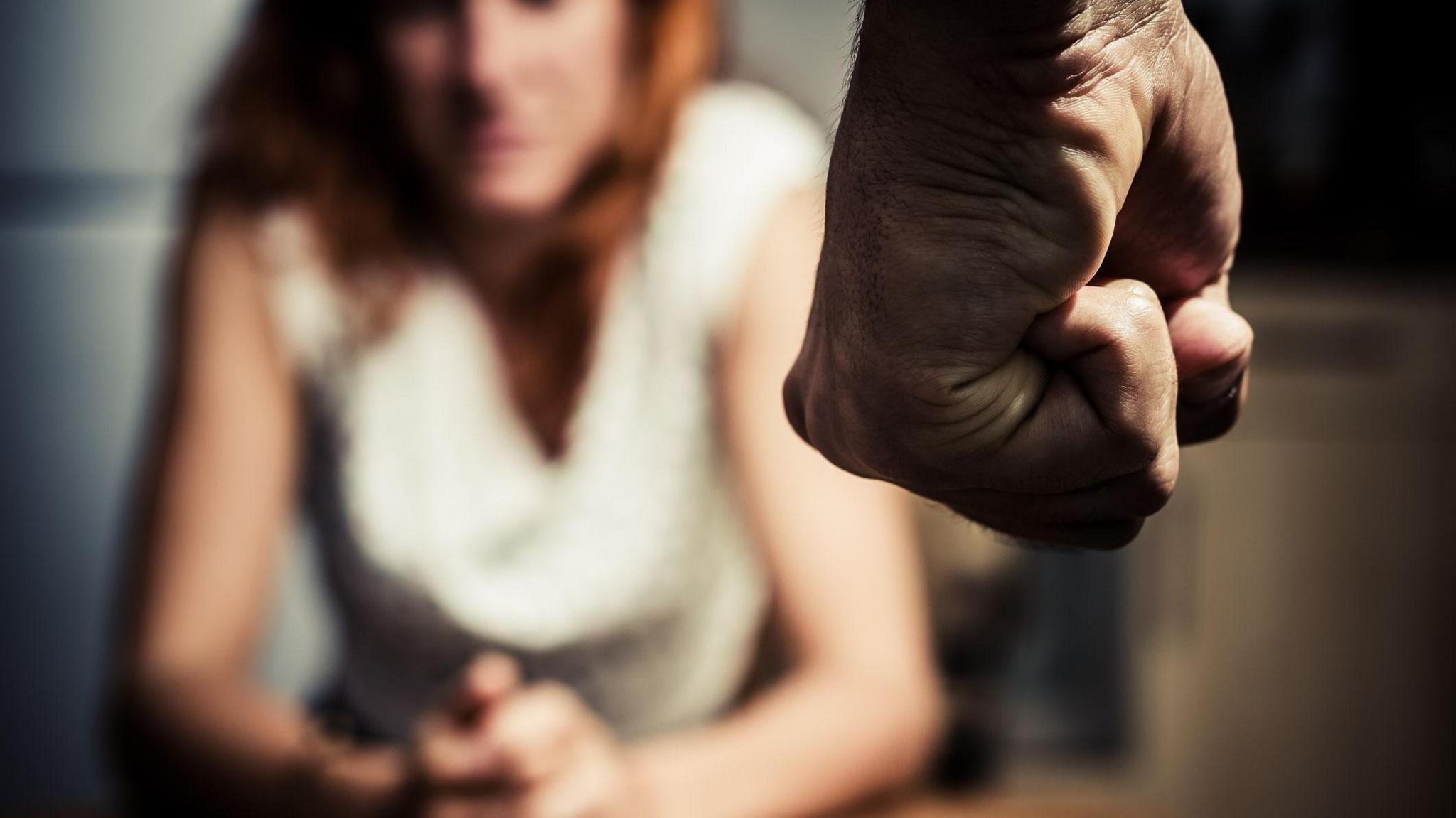 Man's fist clenched in foreground. 
Blurred woman, with red hair wearing a white blouse,  is sitting hunched at a table in the background. 