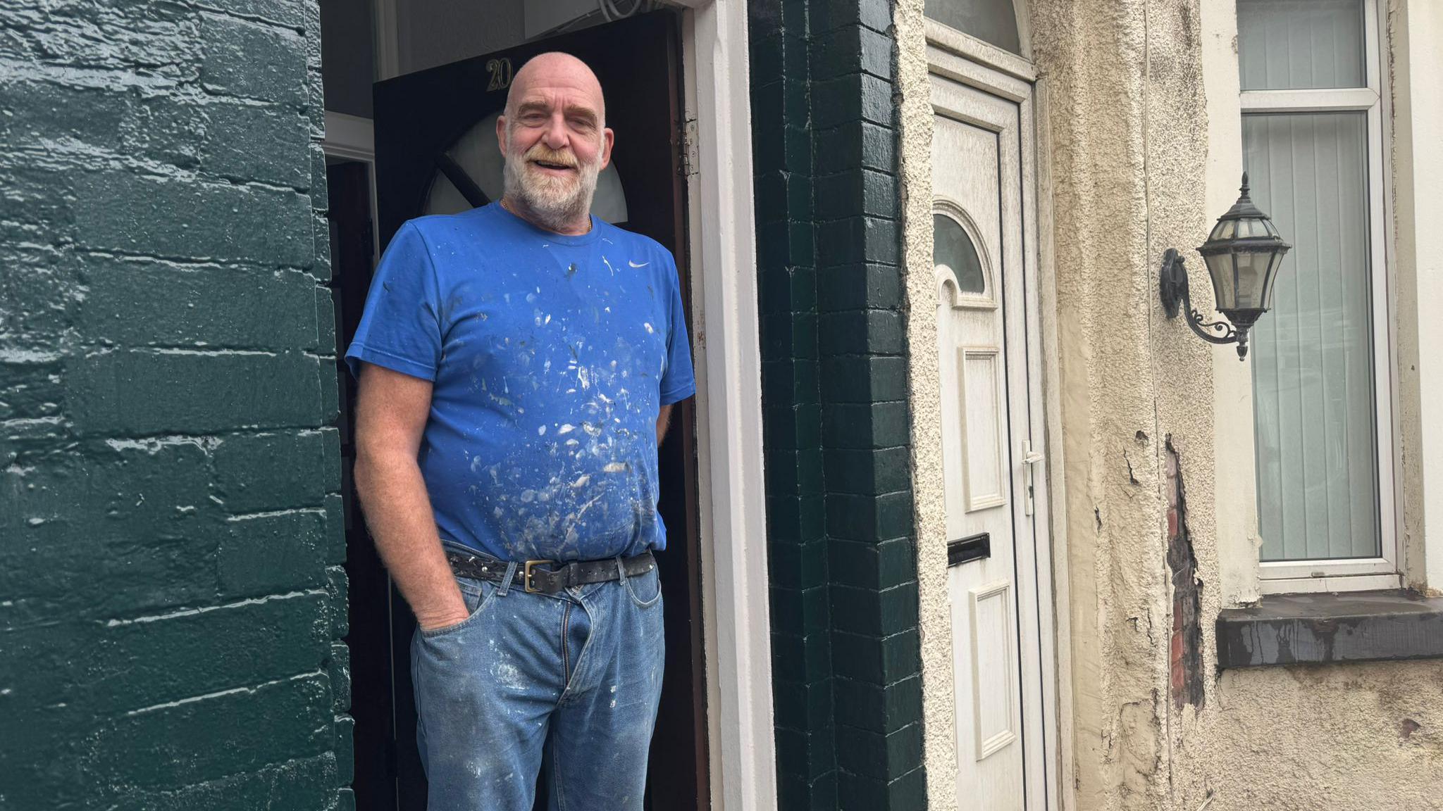 Ken, who is bald and has a grey beard, and wears a blue t-shirt and jeans spattered with white paint from his work, stands in the doorway of a terraced house with the brickwork painted dark green and smiles at the camera. 