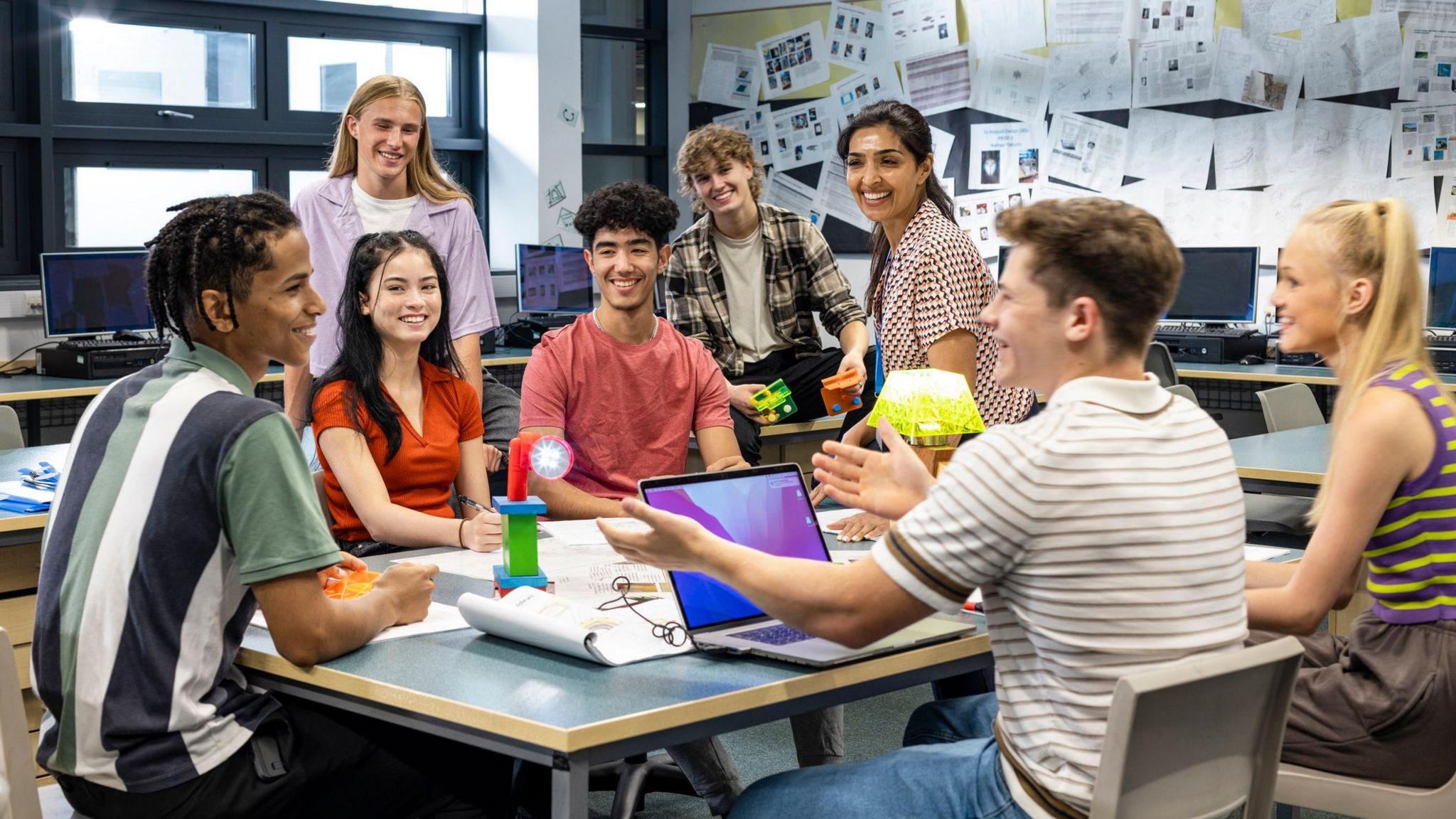 Seven students are sitting around a table. They are with a teacher inside a classroom